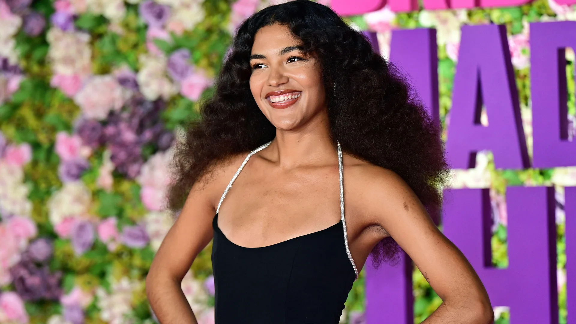 A photo of Olivia Dean wearing a black dress with her hands on hips smiling to someone off the side. Behind her is some purple lettering on a wall and flowers.