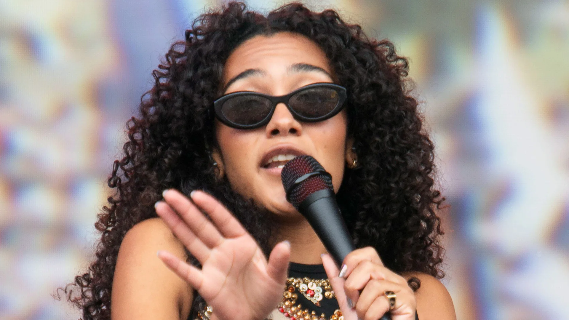 A photo of Olivia Dean singing into a microphone wearing black sunglasses holding her palm outwards
