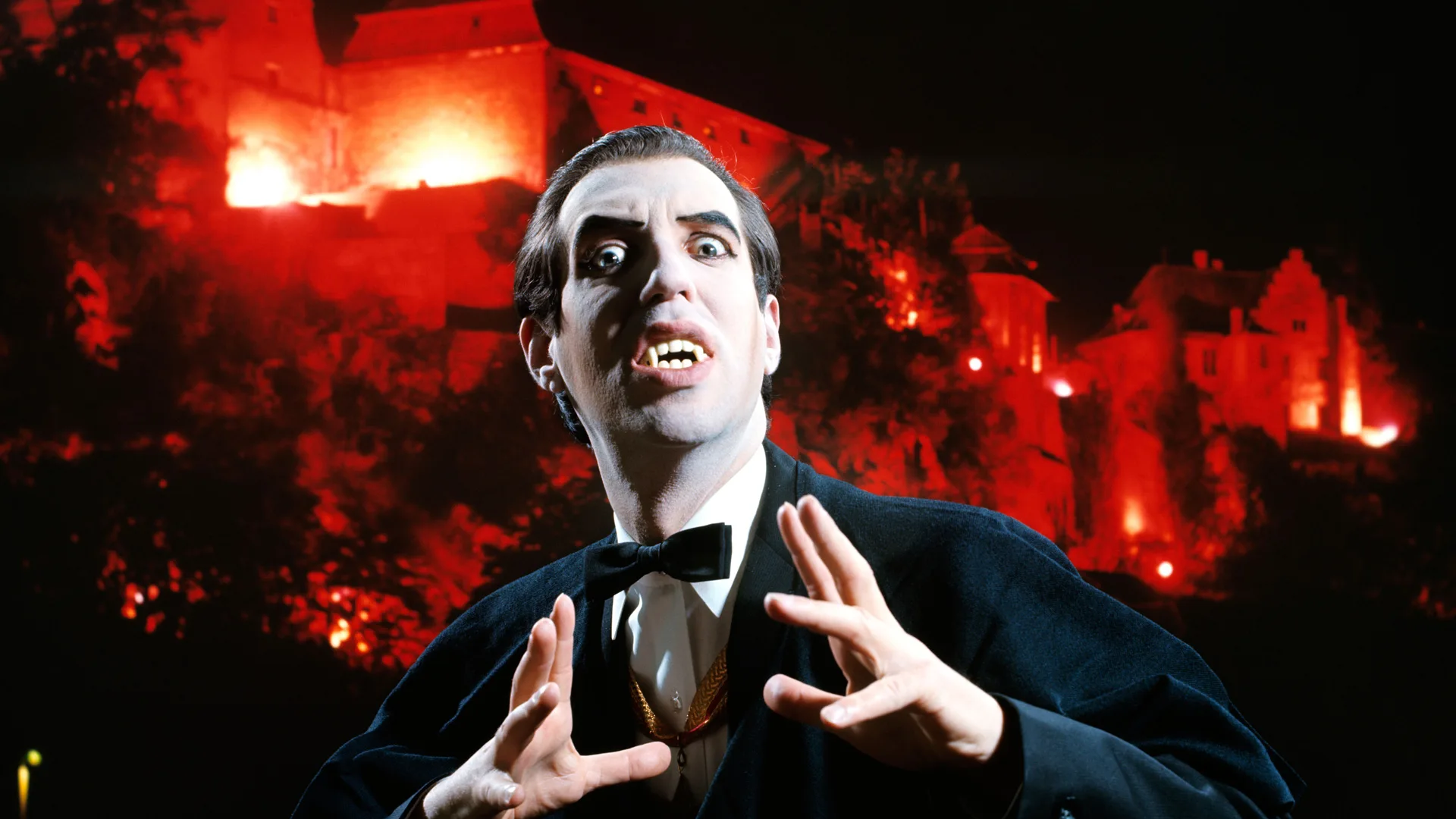 A photo of a man dressed as a vampire holding his hands out with his teeth exposed with a red flaming background behind him.