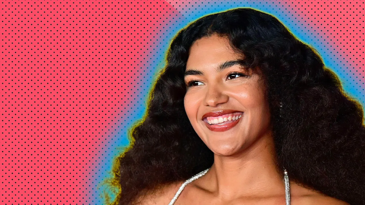 A photo of Olivia Dean wearing a black dress with her hands on hips smiling to someone off the side. She is set against a pink dotted background with a blue halo.