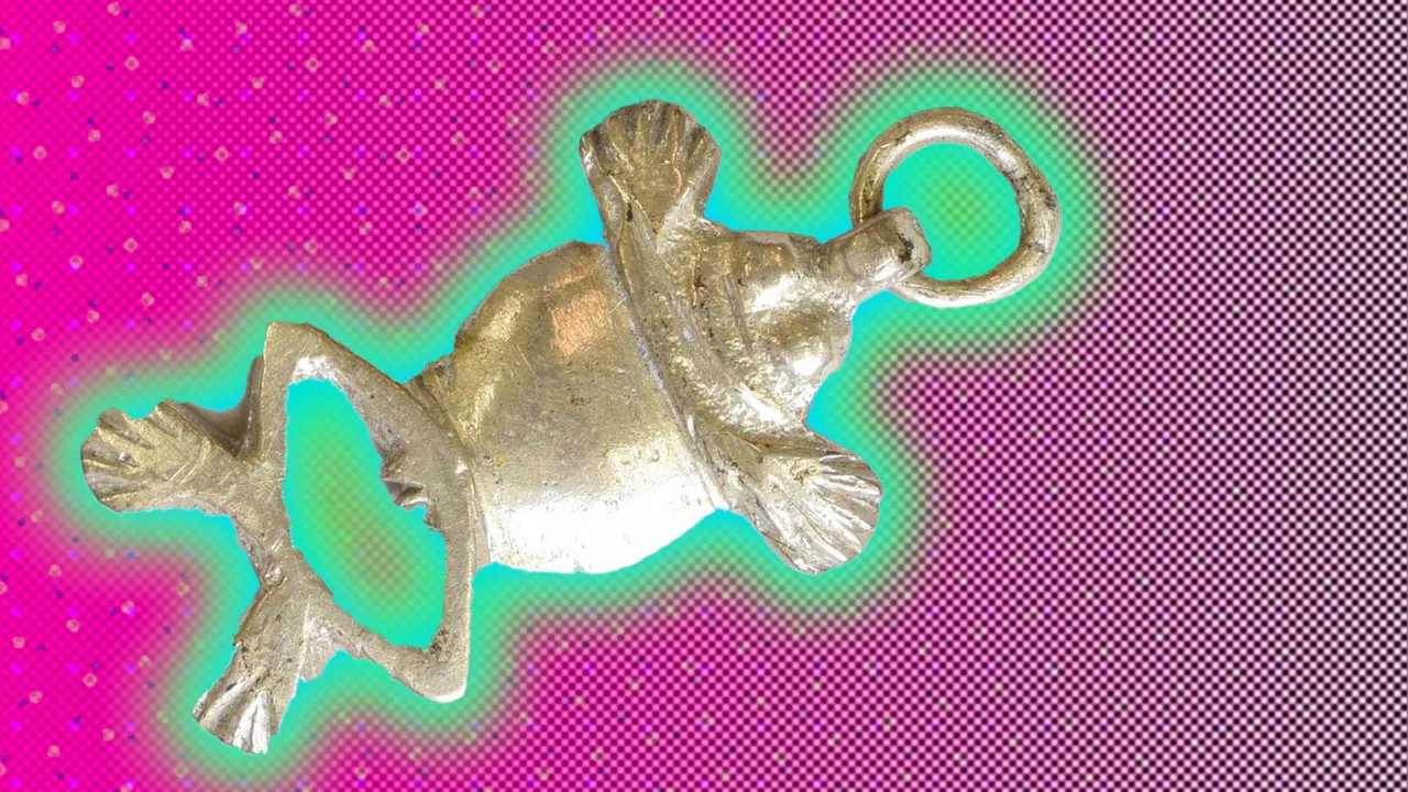 An image of a silver frog charm against a pink background with a green halo.