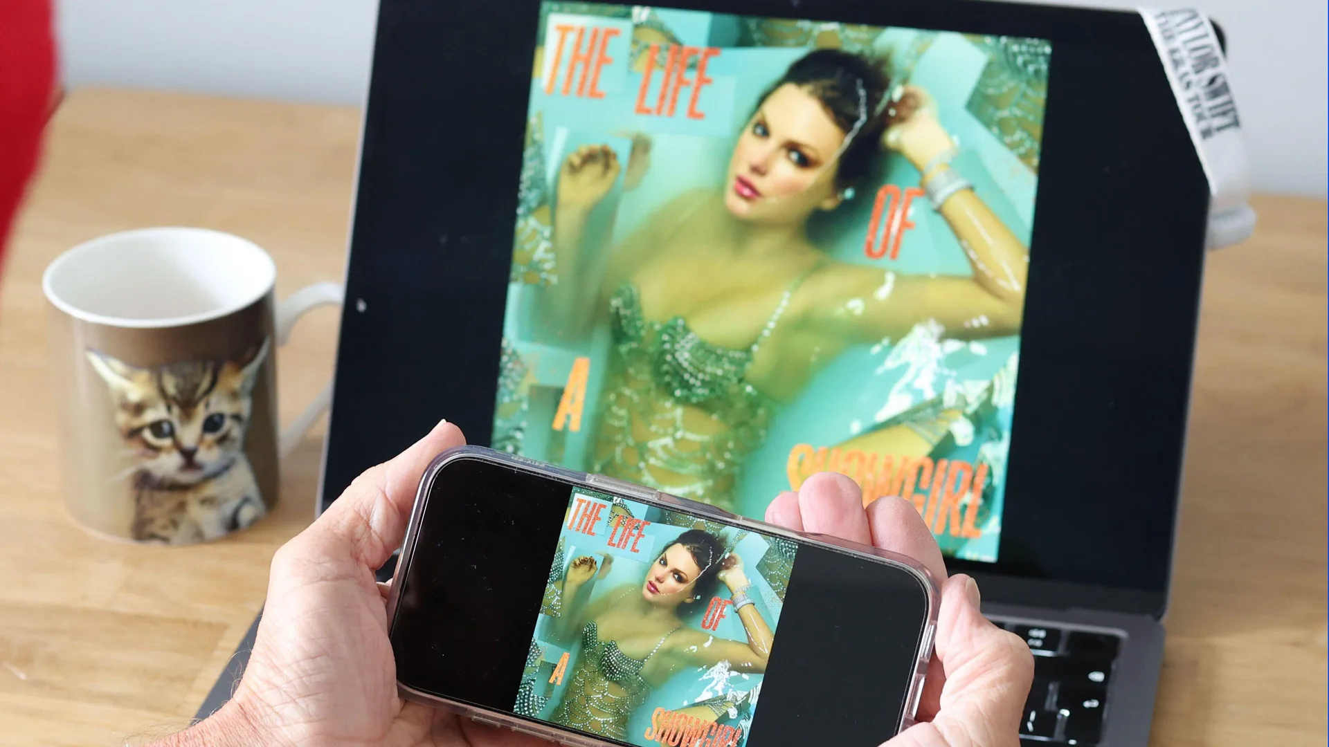 An image of the twelfth studio album by Taylor Swift on a laptop screen showing her wearing a sparkly costume whilst sitting in water with her hand up to her head. Someone is holding a phone near the laptop screen taking a picture of it.