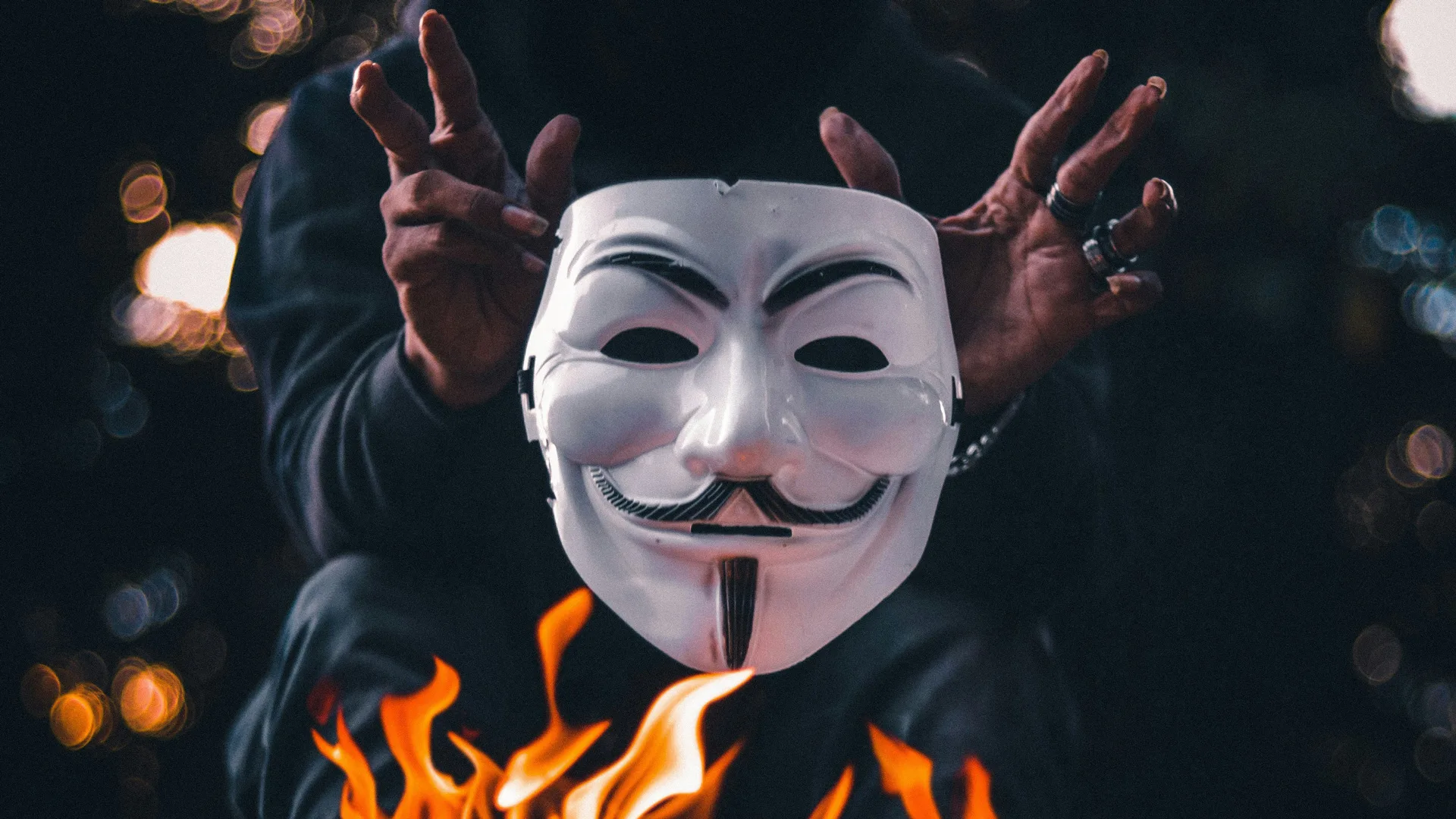 A photo of a black and white mask with a moustache and grin being held above flames by a pair of hands against a dark background.