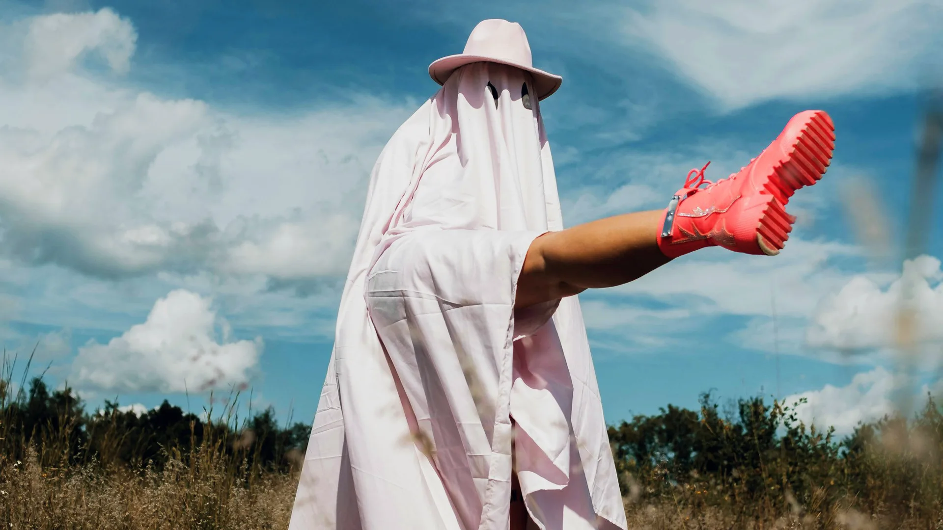A photo of a person wearing a white sheet like a ghost with a white hat on top and they are kicking their leg out wearing a pink boot. They are stood in a field of grass and trees with a blue cloudy sky behind.