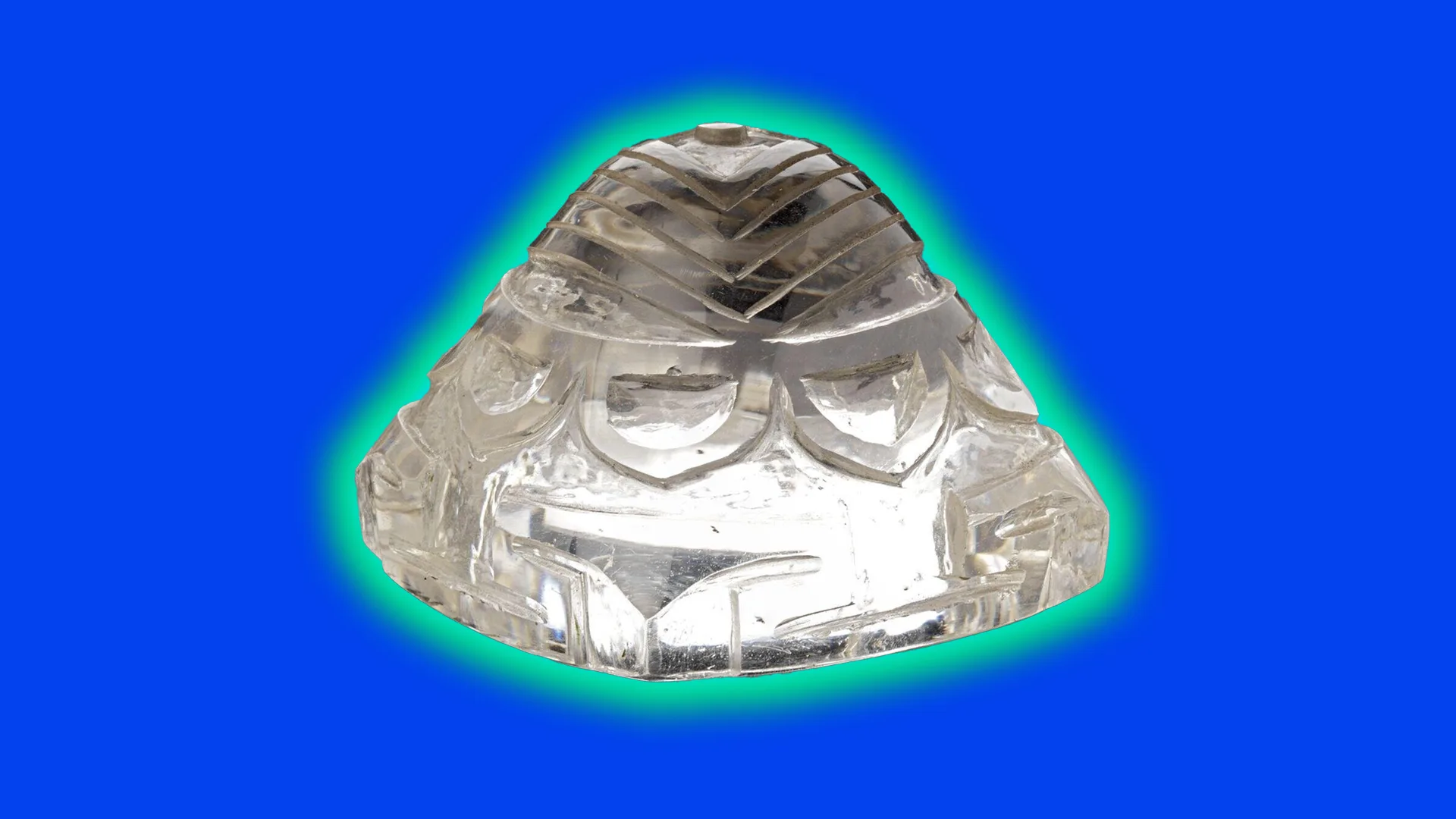 A crystal clear charm shaped like a mountain with carvings around it set against a blue background with a blue green halo.