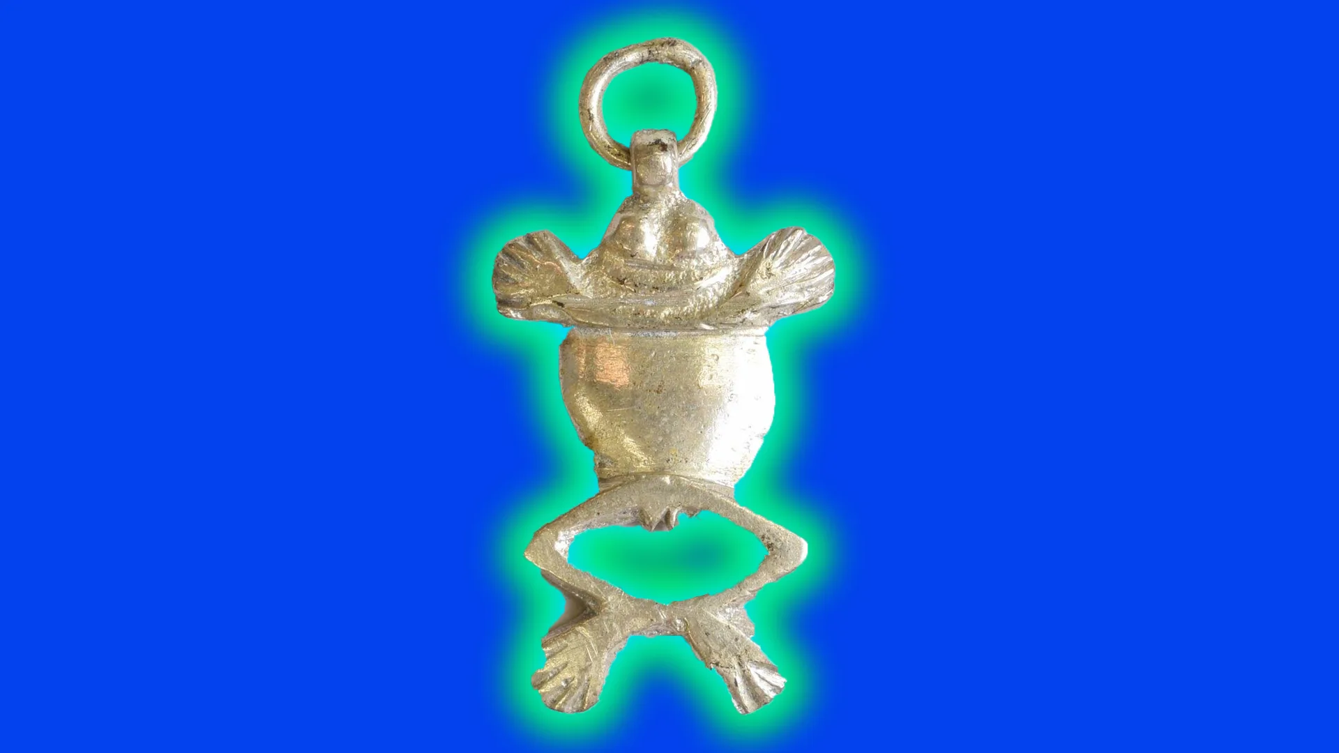An image of a frog charm made out of silver showing a birds eye view of a frog with its front hands stretched out and bag legs in a bow pose. It is set against a blue background with a green halo.