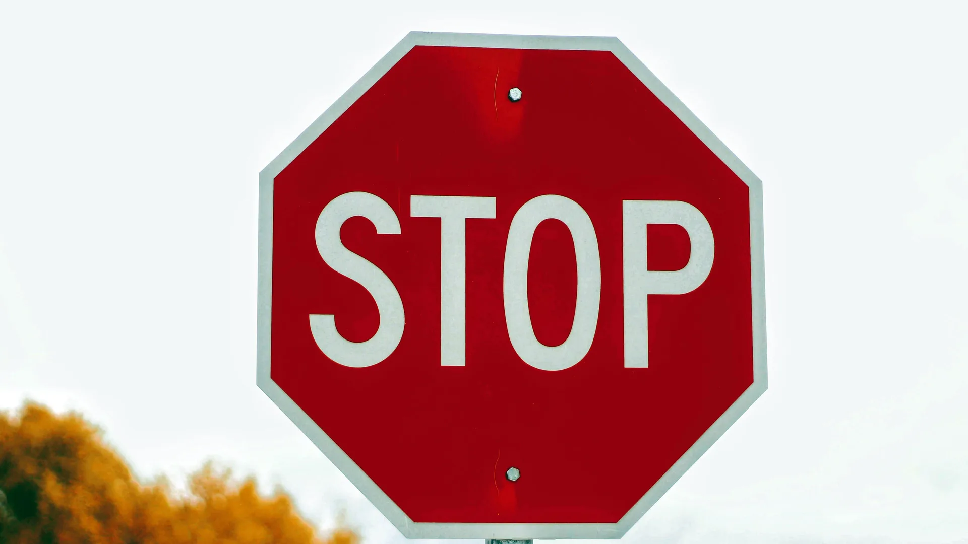 An image of a red and white stop sign against a light sky background with some grass showing in the bottom left.