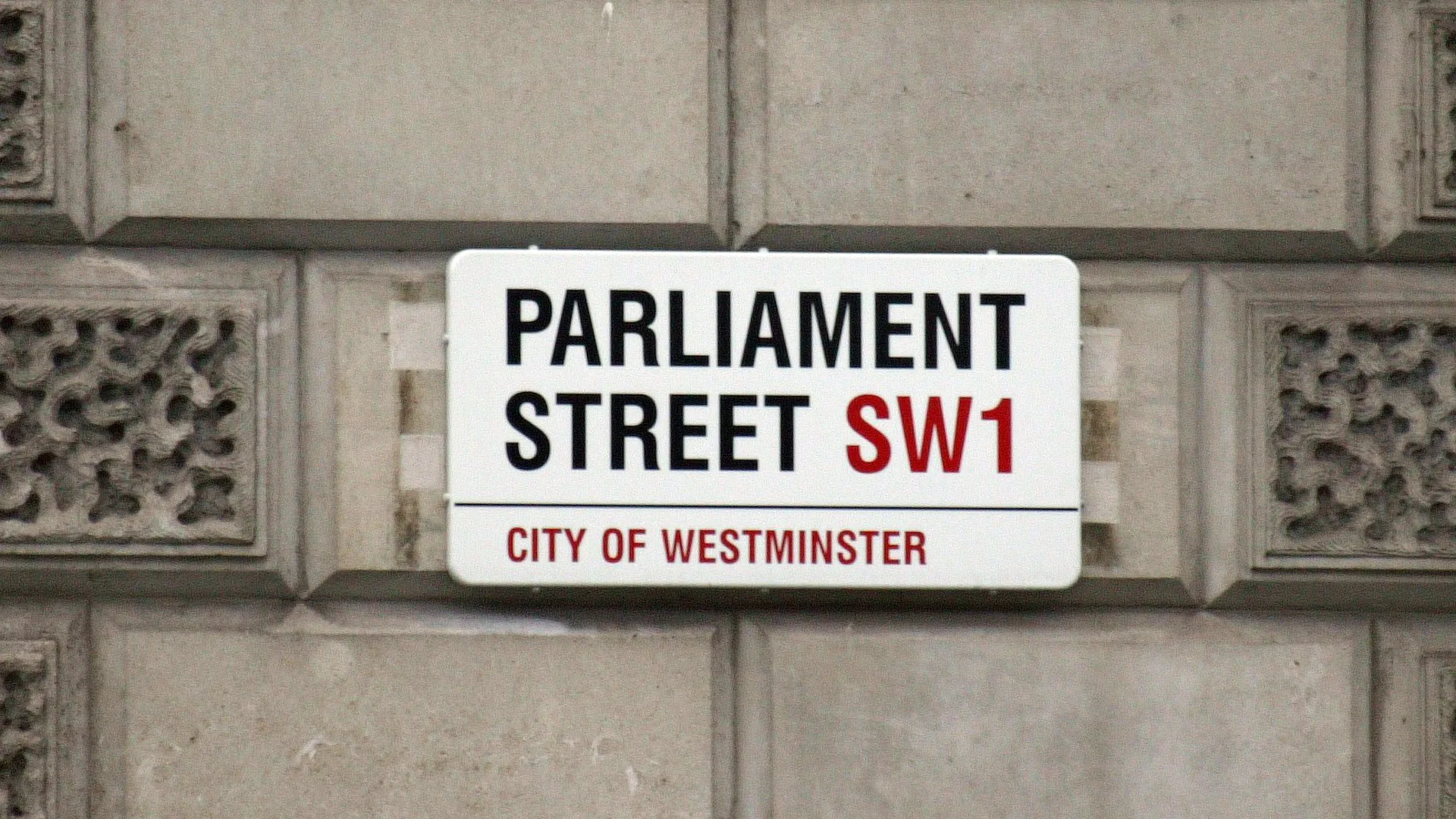 A black and white sign that says PARLIAMENT STREET SW1 - CITY OF WESTMINSTER is red text on a brick wall.