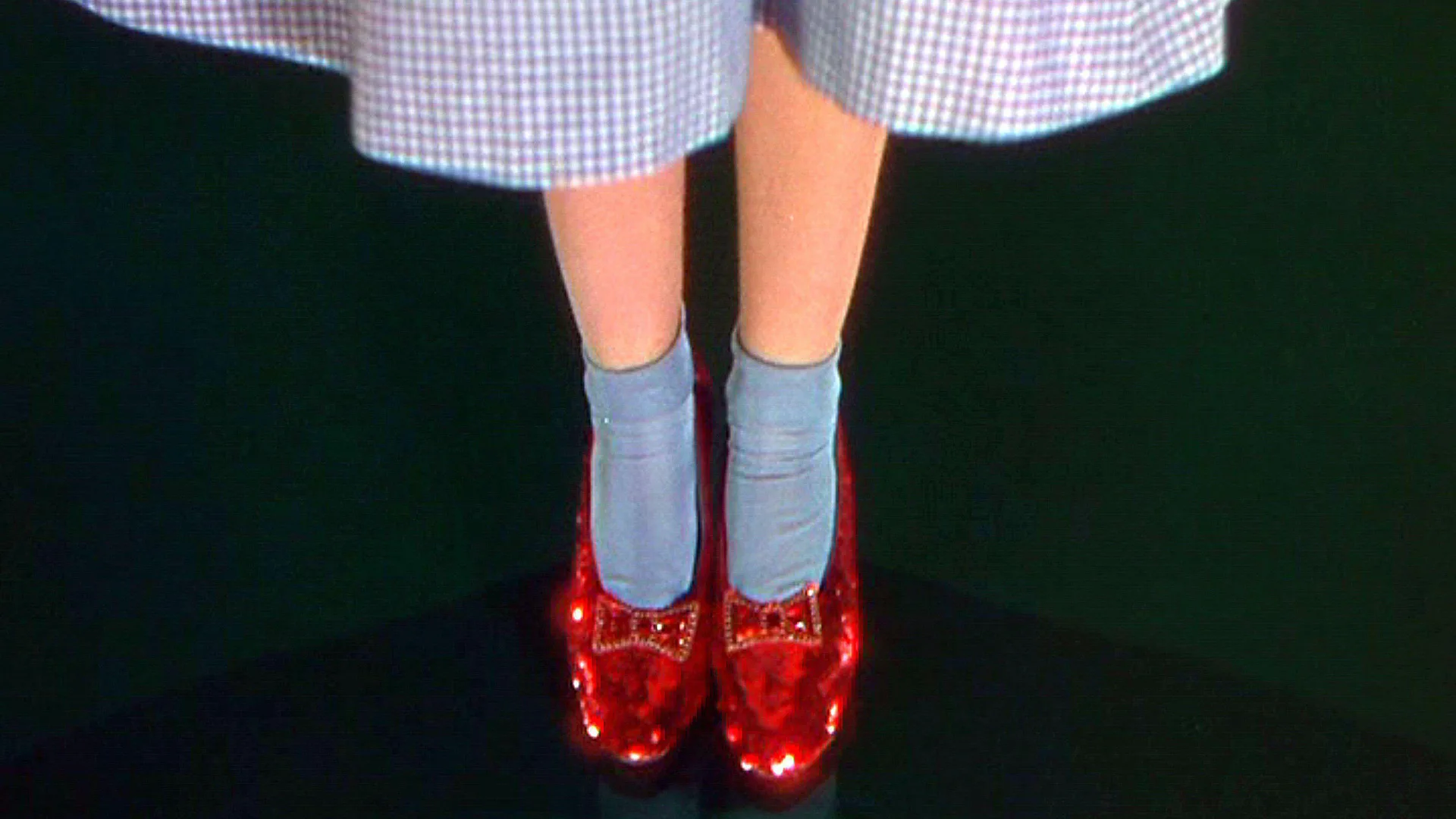 A still from the movie The Wizard of Oz showing Dorothy's feet wearing the ruby red slippers and blue socks with her blue gingham dress showing. She is stood on a black floor.