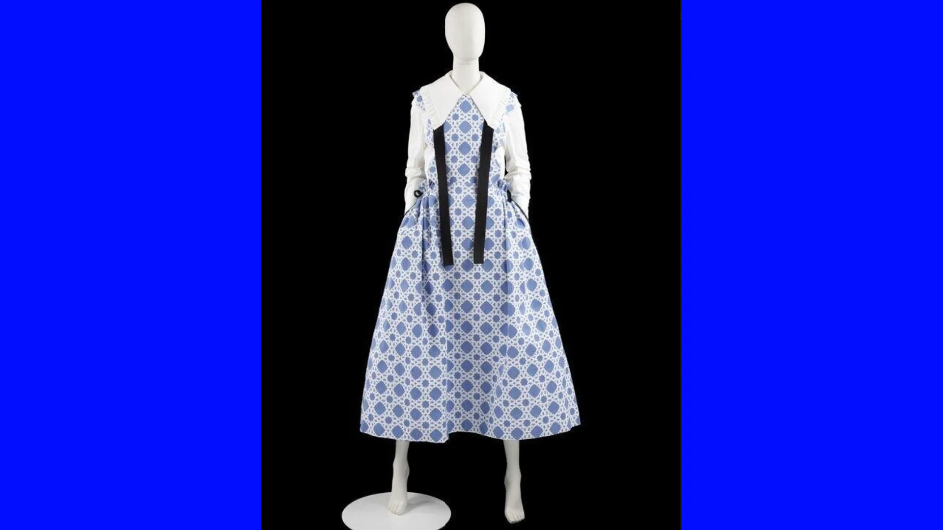 A photo of a blue woven moon jar patterned dress with white peter pan collar and sleeves and black tie ribbons hanging down the front. It is on a white mannequin with one foot on a podium set against a black background with blue borders either side.