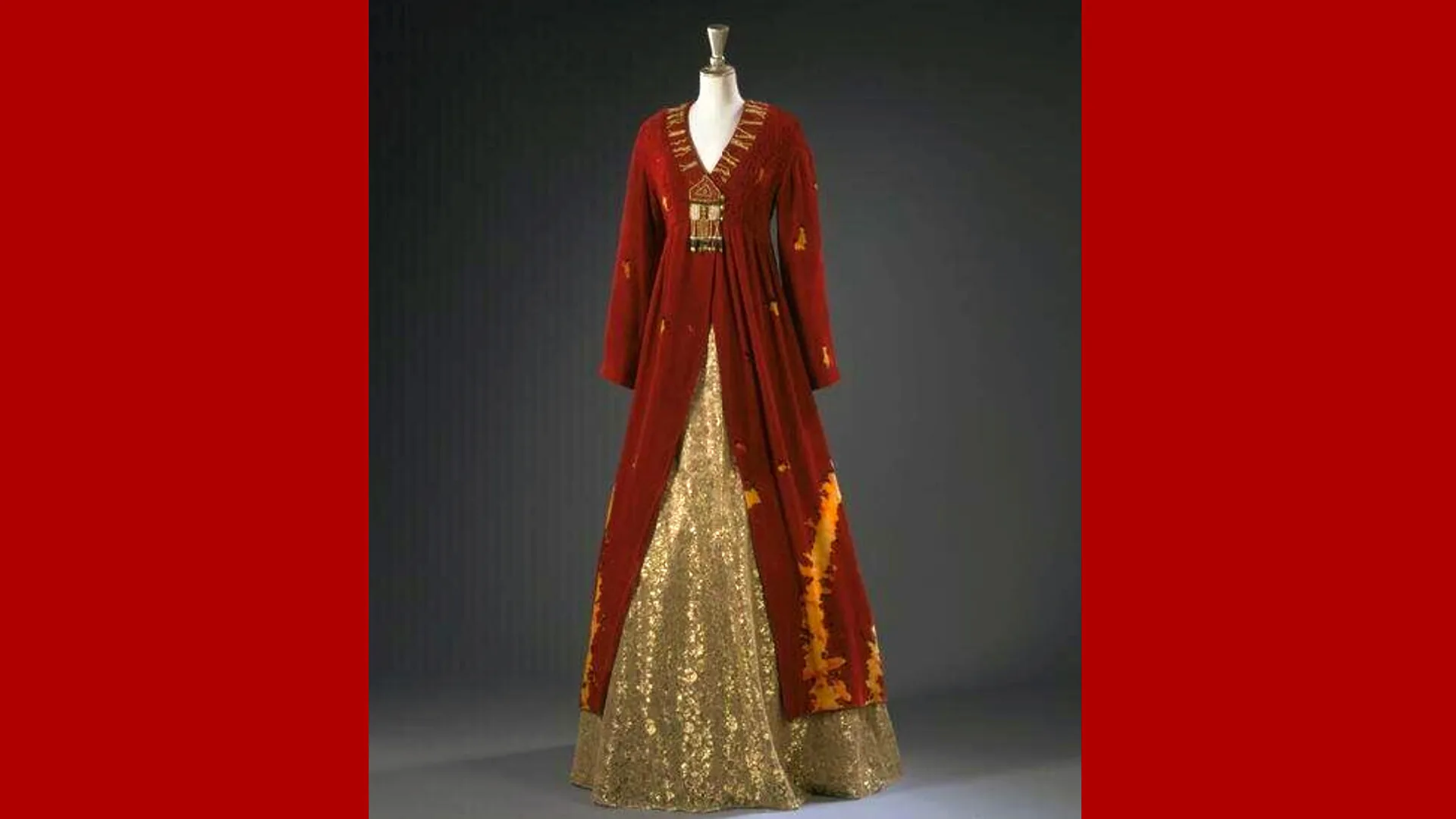 A photo of a designer dress in red silk velvet and gold lace and tulle set against a grey background with red borders either side.