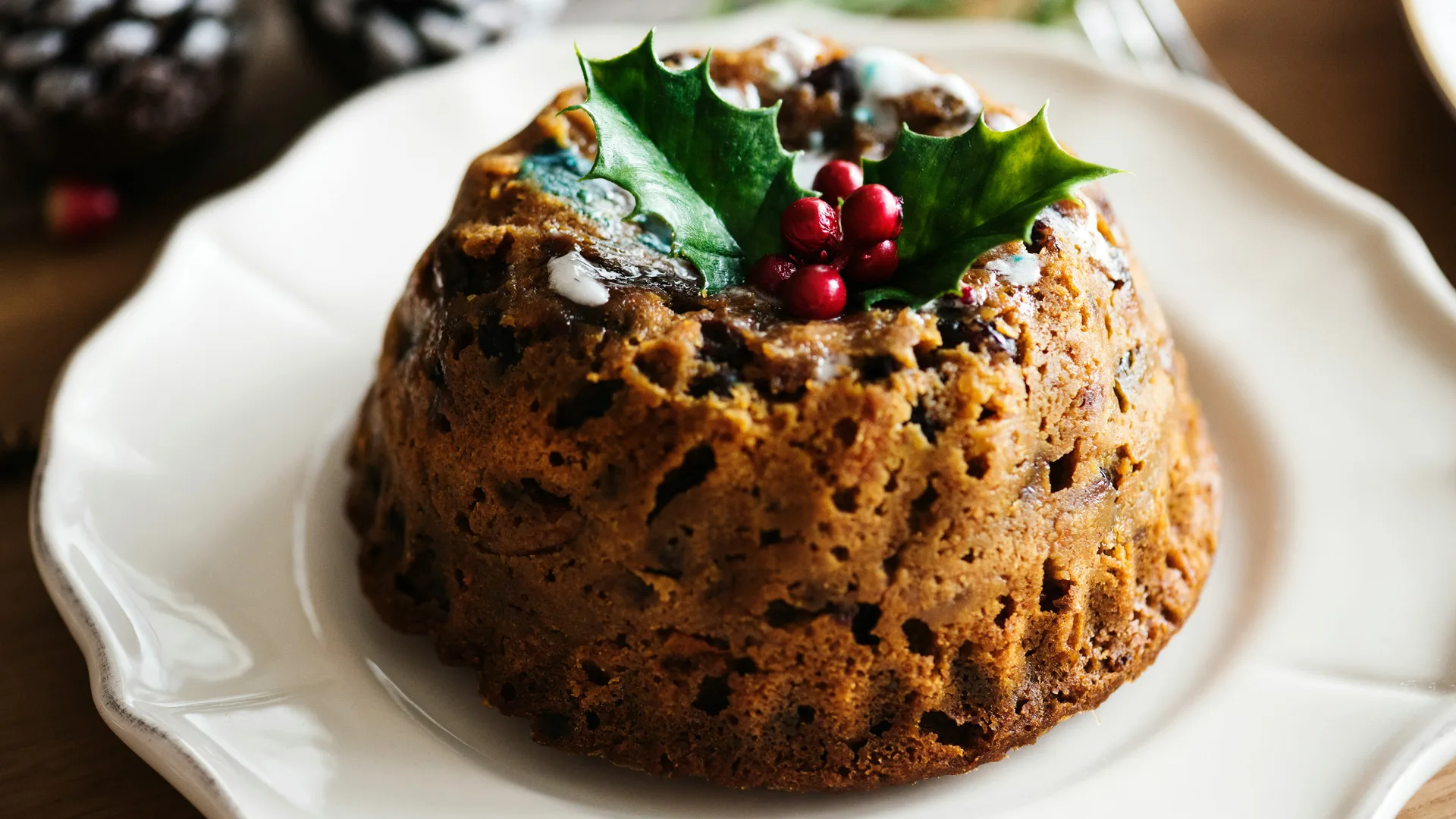 A photo of a Christmas pudding with holly and berries on top sat on a white china plate.