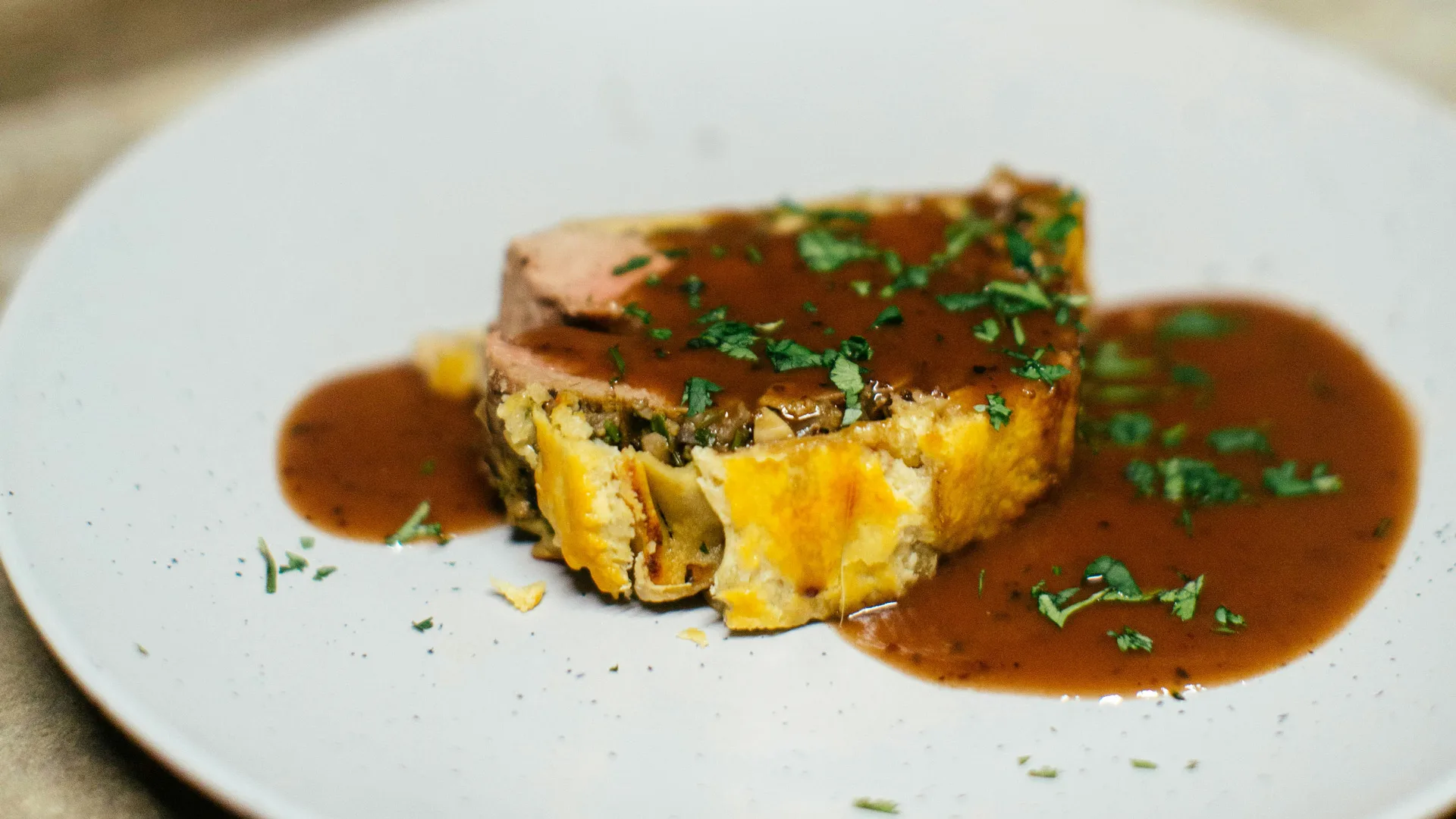 A photo of a slice of beef wellington on a white plate covered in gravy and herbs.