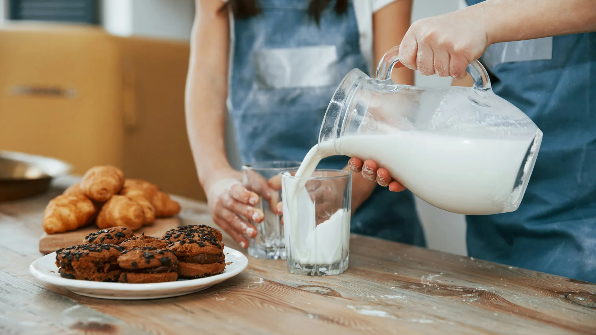 A photo of two people stood by a table with croissants and cookies on a plate, pouring milk from a jug into a glass.