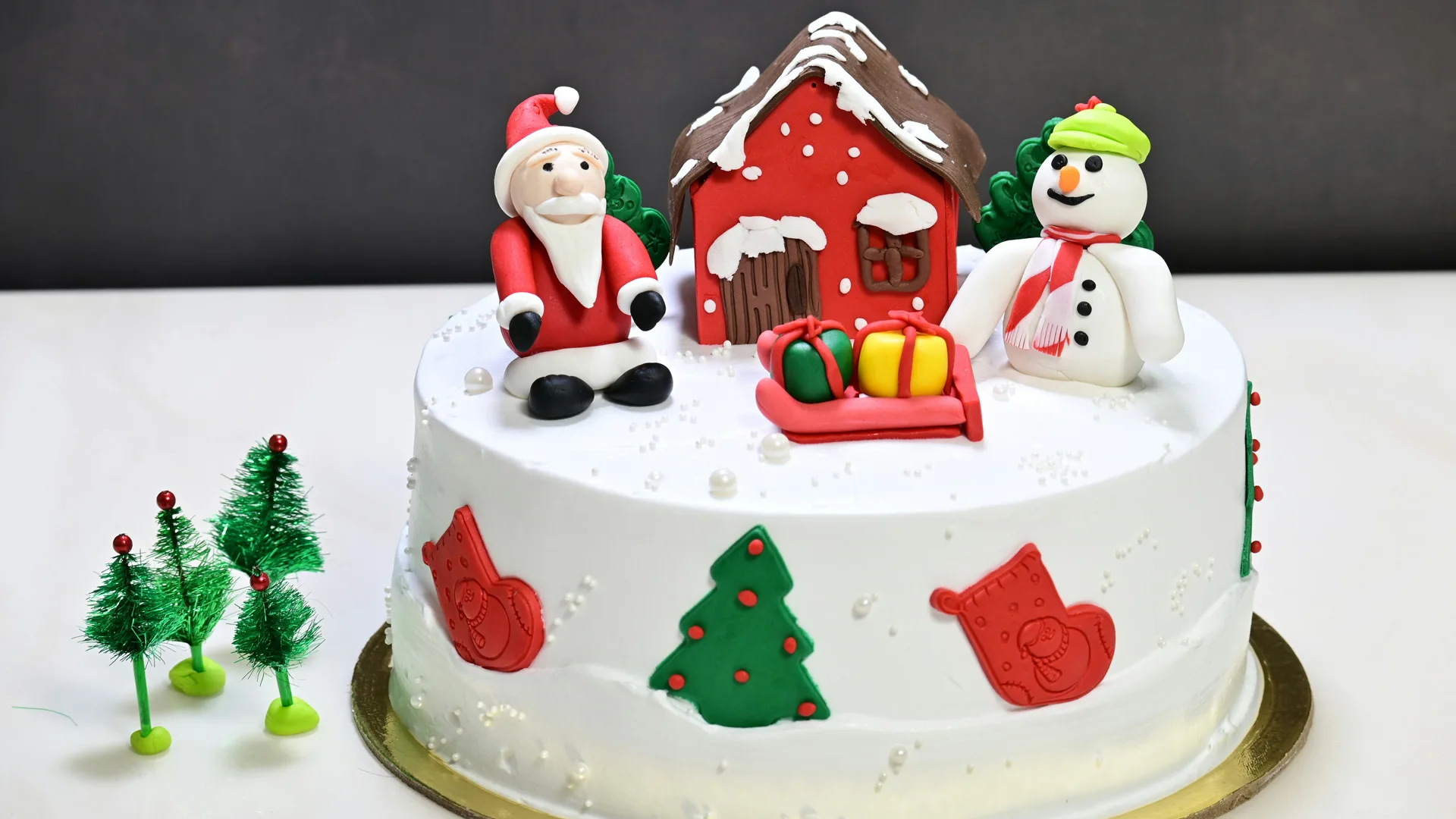 A photo of a white iced Christmas cake with an icing scene of Santa, a snowman, house and presents on the top and around the side are red stockings and green christmas trees.