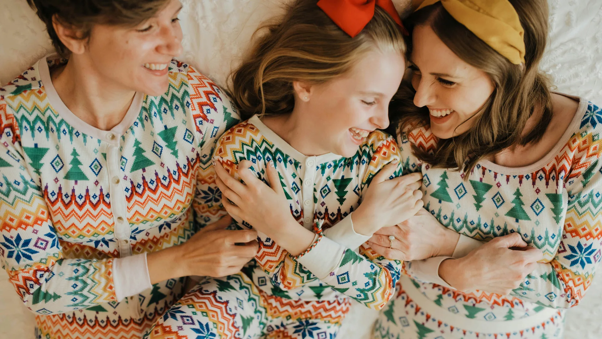 A family of three lying on a bed wearing matching christmas pyjamas all laughing and holding one anothers arms.