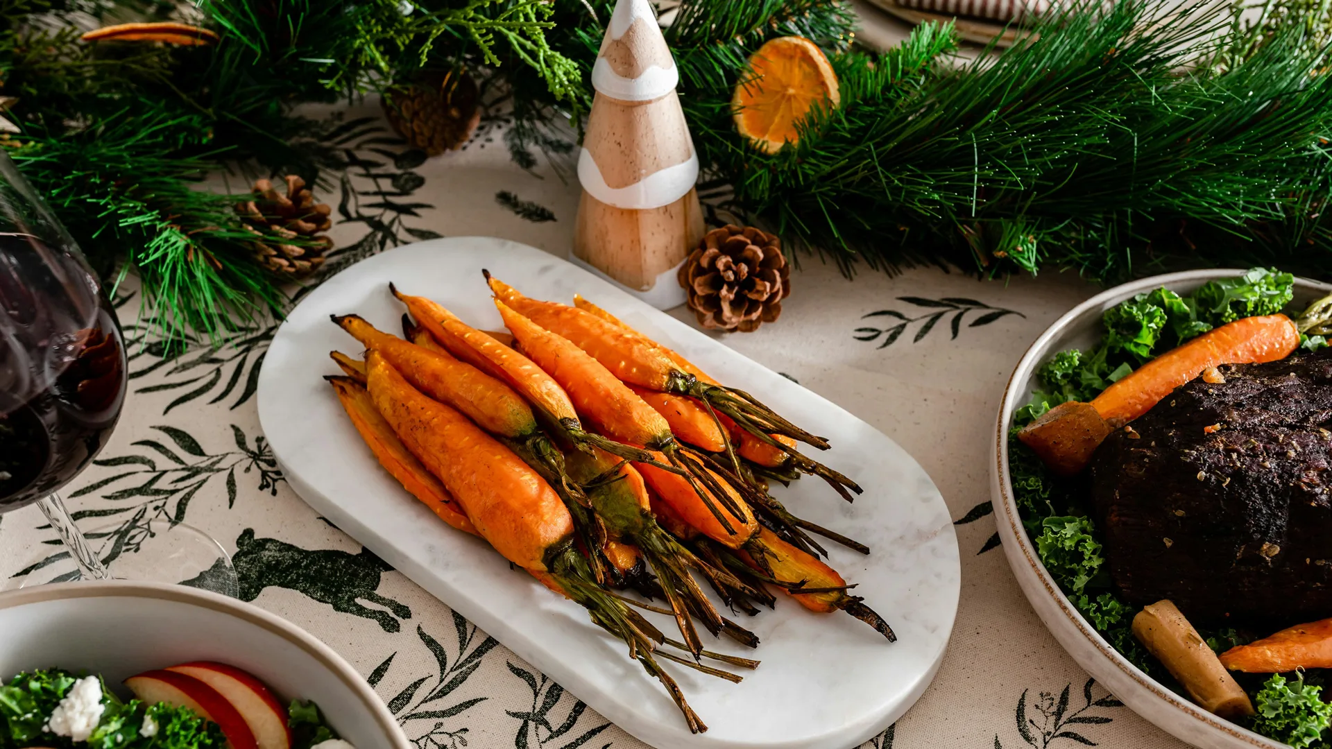 An image of roasted carrots on a white plate surrounded by festive decor such as a wooden christmas tree with white snow and a green wreath with dried oranges.