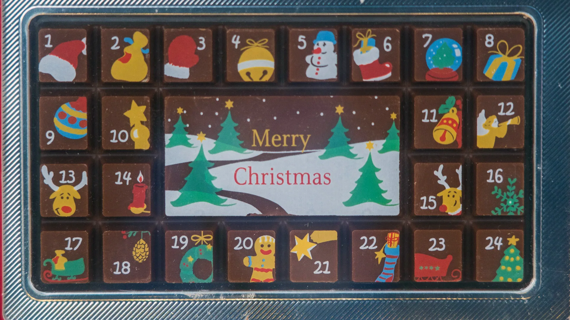 A photo of a box of chocolates with different Christmas-related pictures on them and in the middle a sign that says "Merry Christmas" with a snowy field scene with christmas trees.
