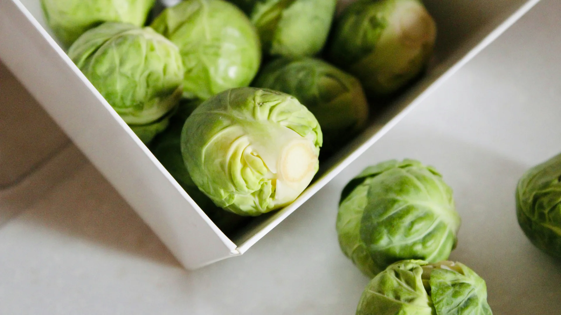 A photo of some brussel sprouts in a white square dish with two sprouts outside of it to the side on a white table.