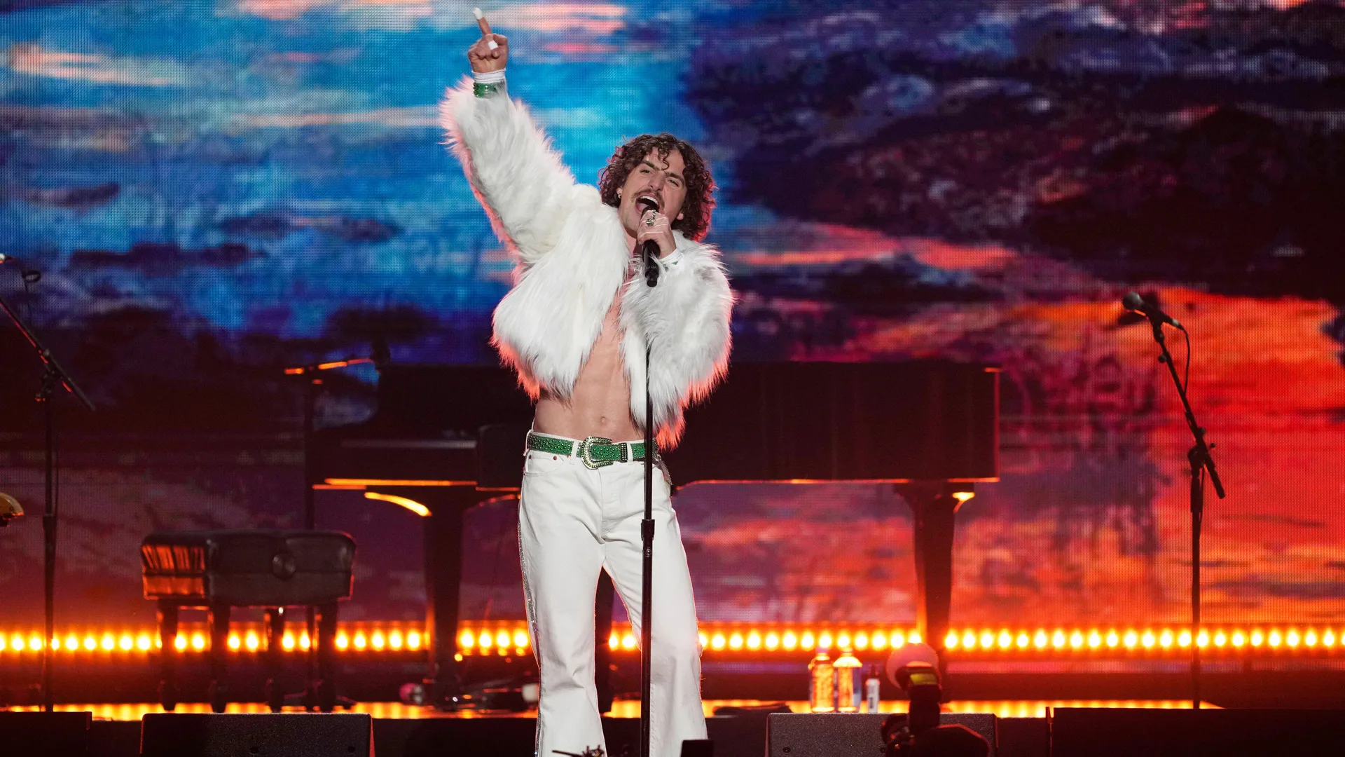 A photo of Benson Boone singing on stage wearing a white jacket and jeans with his arm up in the air set against a painted sunset background.