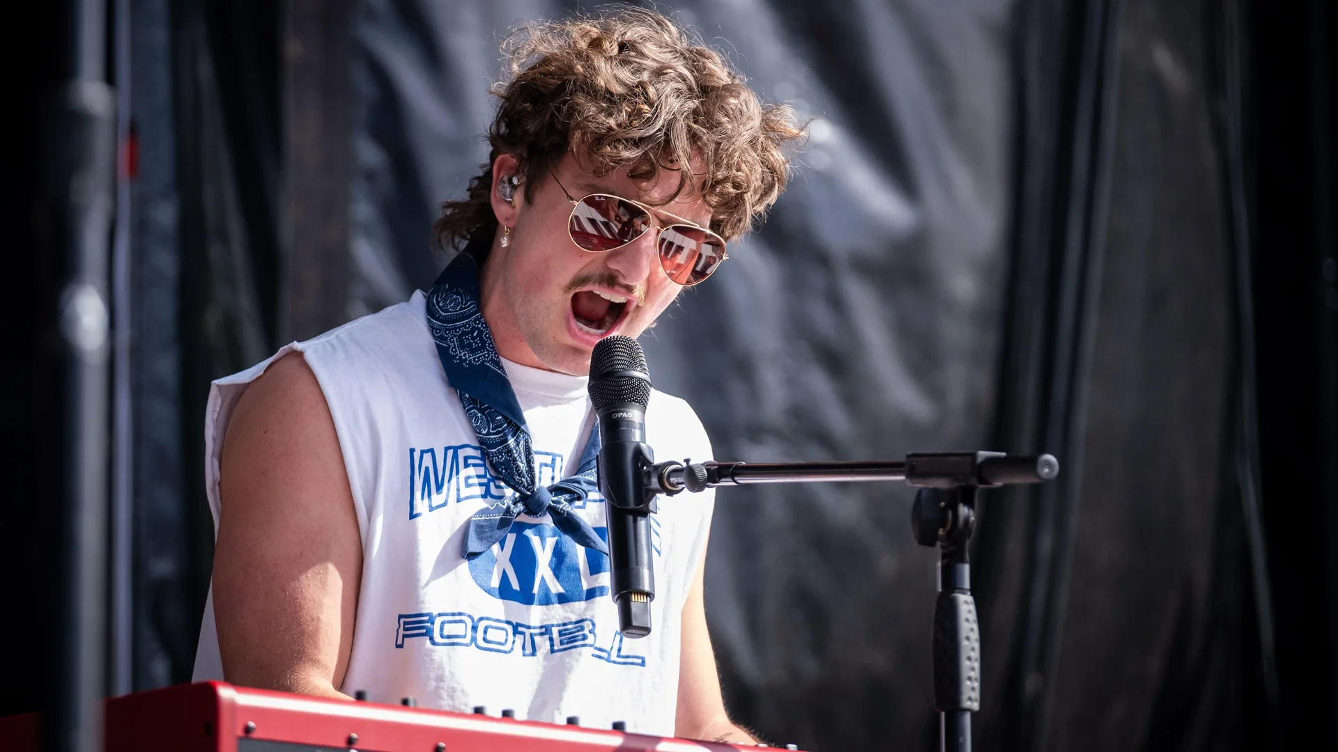 A photo of Benson Boone singing into a mic wearing a white tshirt set against a black background.