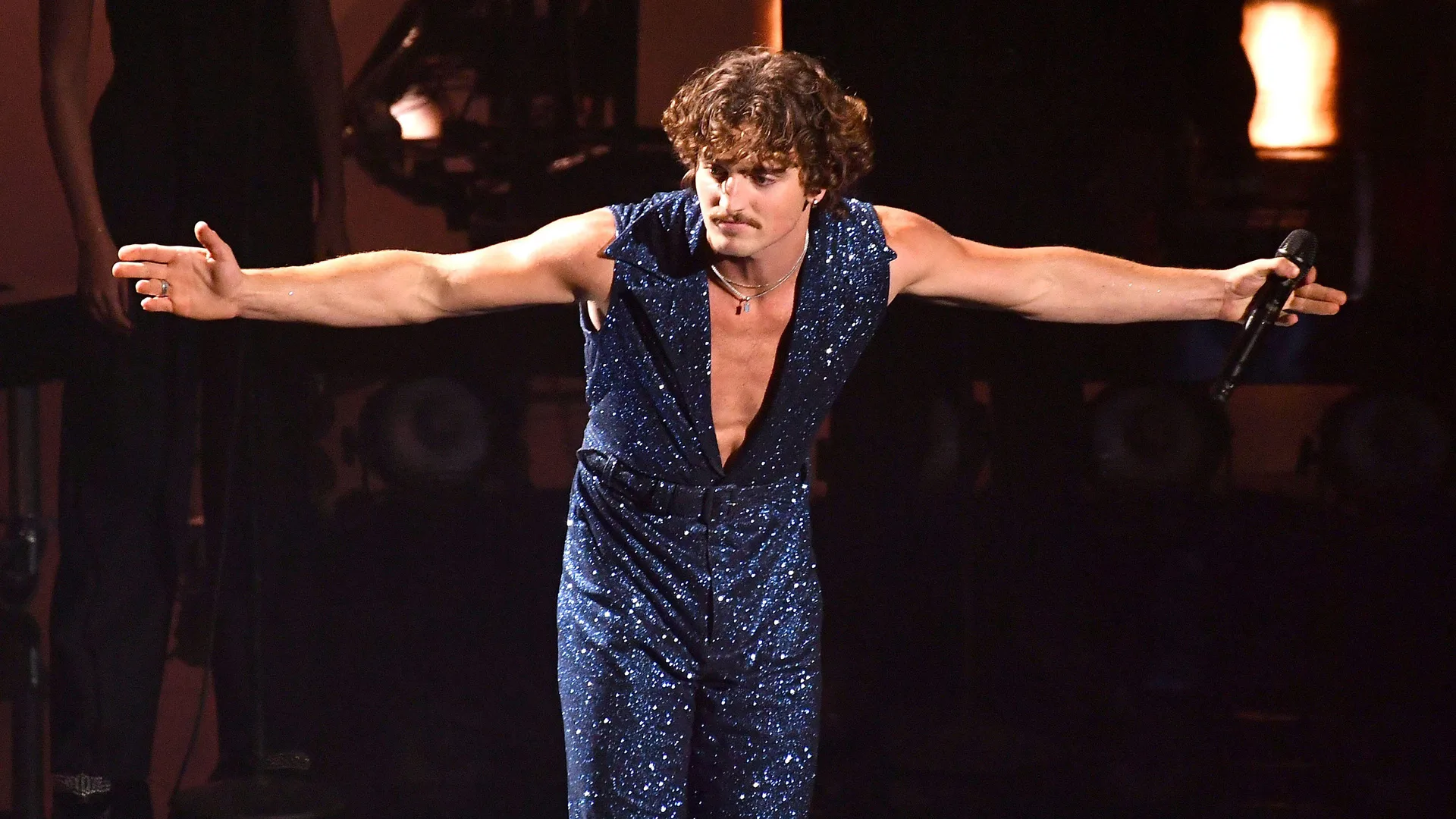 A photo of Benson Boone stood on stage with arms stretched out wearing a blue sparkly suit.