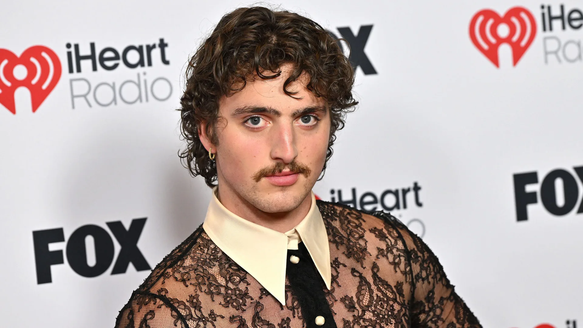 A photo of Benson Boone showing him looking right at the camera wearing a lace shirt with a white collar and black tie set against a banner with FOX and iHEART RADIO across it.