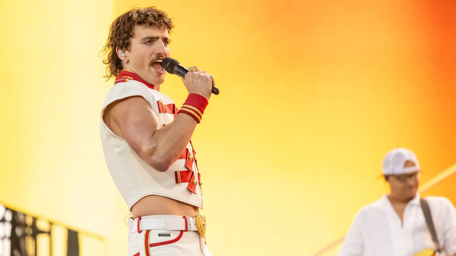 A photo of Benson Boone singing on stage with a mic in a white and red military outfit with a yellow and orange background.
