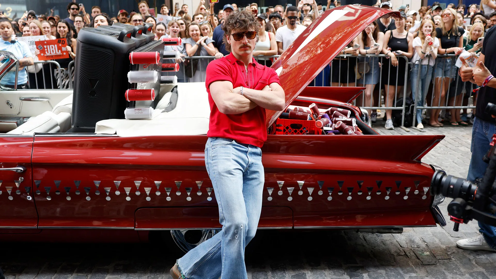 A photo of Benson Boone stood arms folded against a red car - he's wearing blue jeans and a red tshirt and sunglasses.