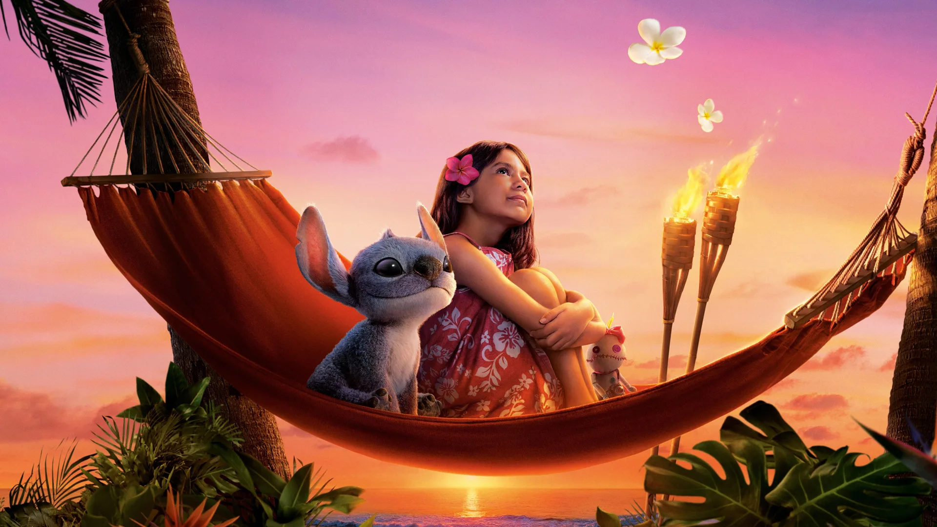 A still from the live action Disney film Lilo and Stitch showing Lilo and Stitch sat in a hammock looking up at the pink sunset sky with two flames beside them and the sun setting on the ocean in the distance.