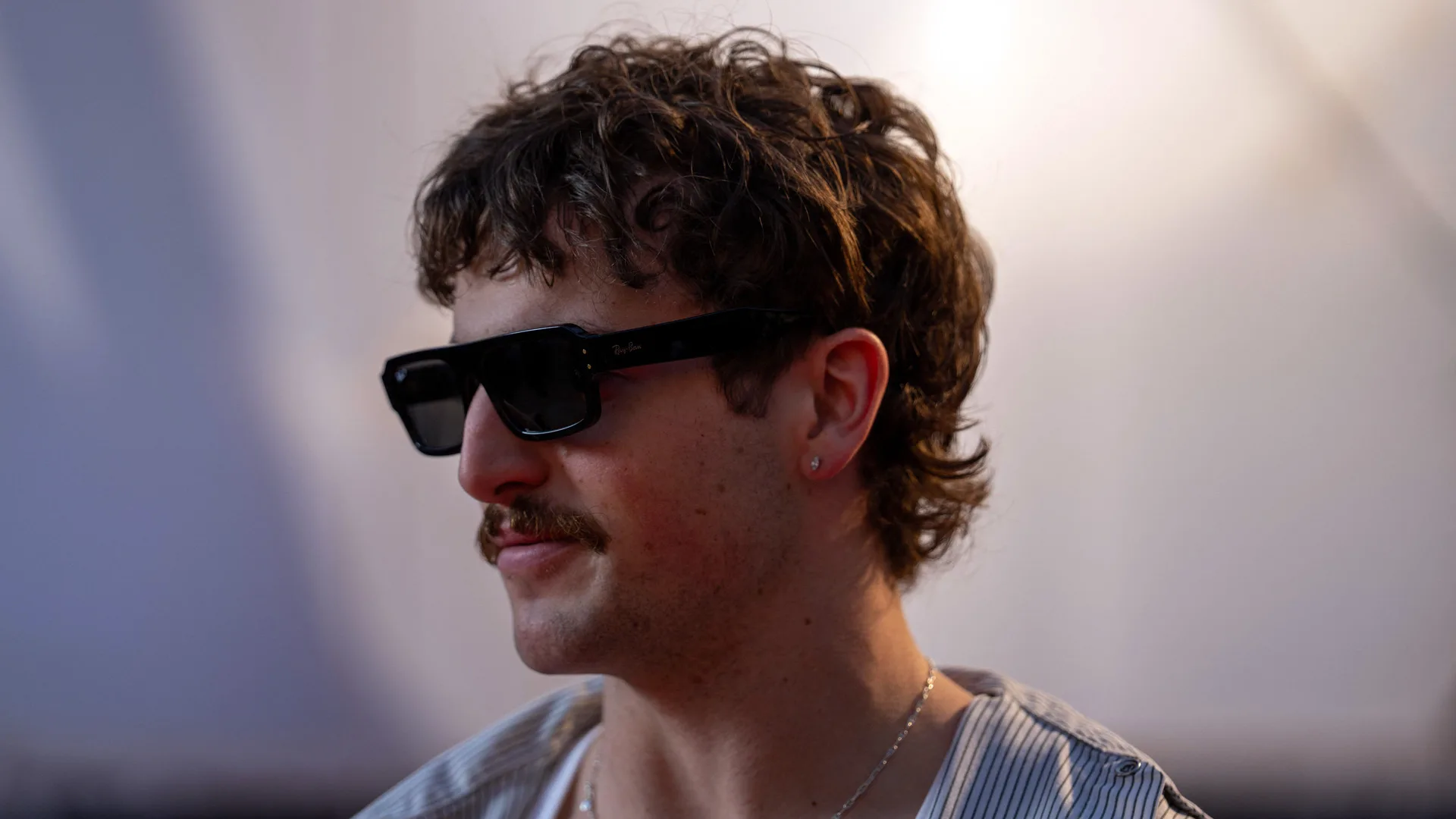 A photo of Benson Boone stood in sunglasses looking to the side with a soft light behind.