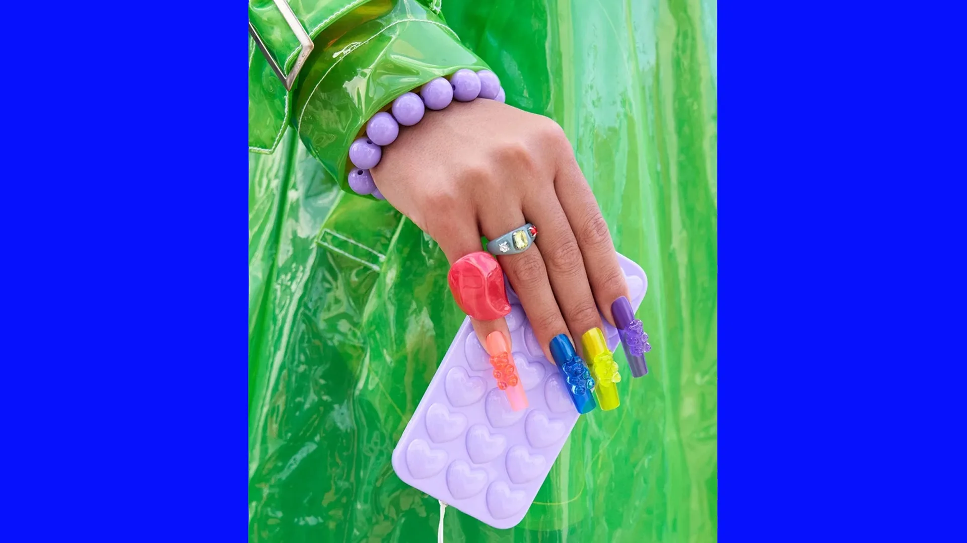 A photo of a hand with brightly coloured long fake nails holding a lilac plastic phone case with heart moulds. The person is wearing a green shiny raincoat with a purple beaded bracelet around their wrist. The image is bordered with blue either side.