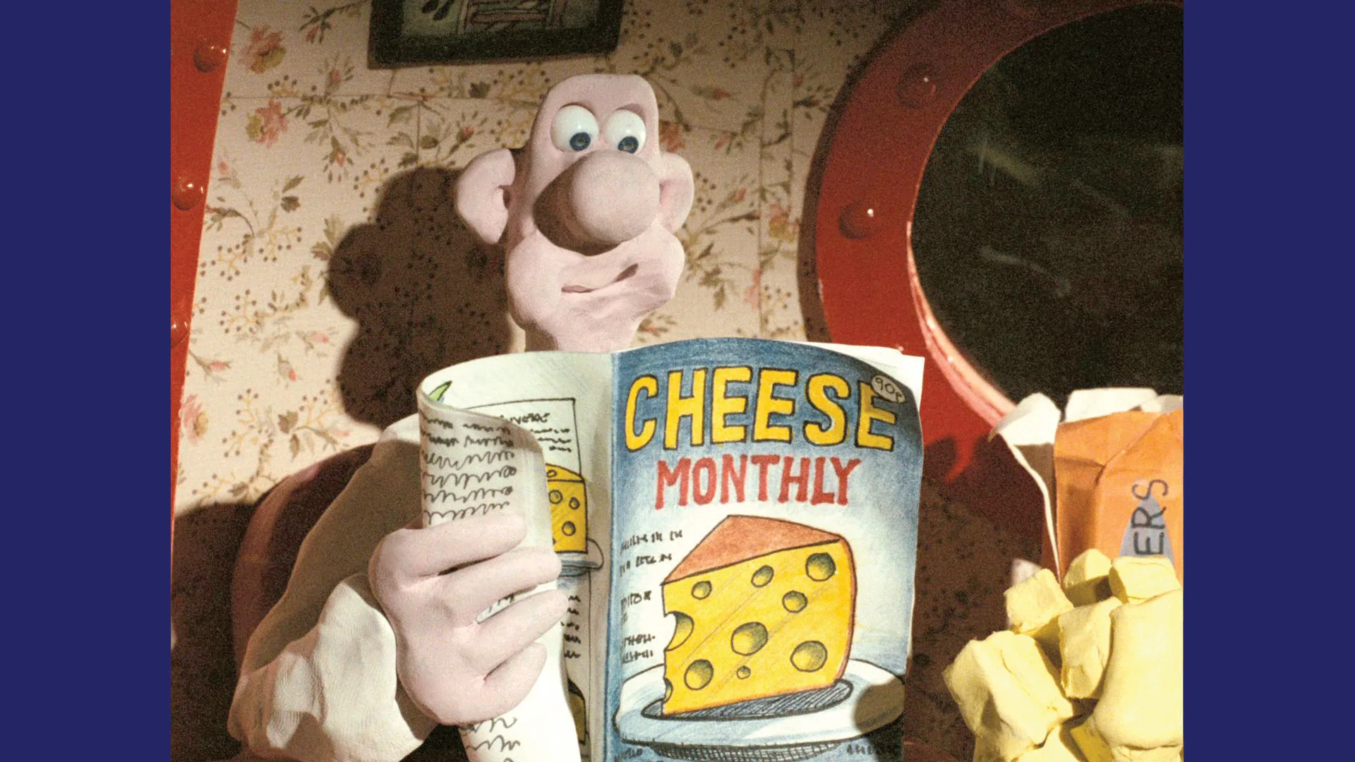 An image of Wallace from A Grand Day Out reading a Cheese Monthly magazine with a holed cheese on the front cover. He is mid-eating sat against a wallpapered background with a red porthole window next to him.