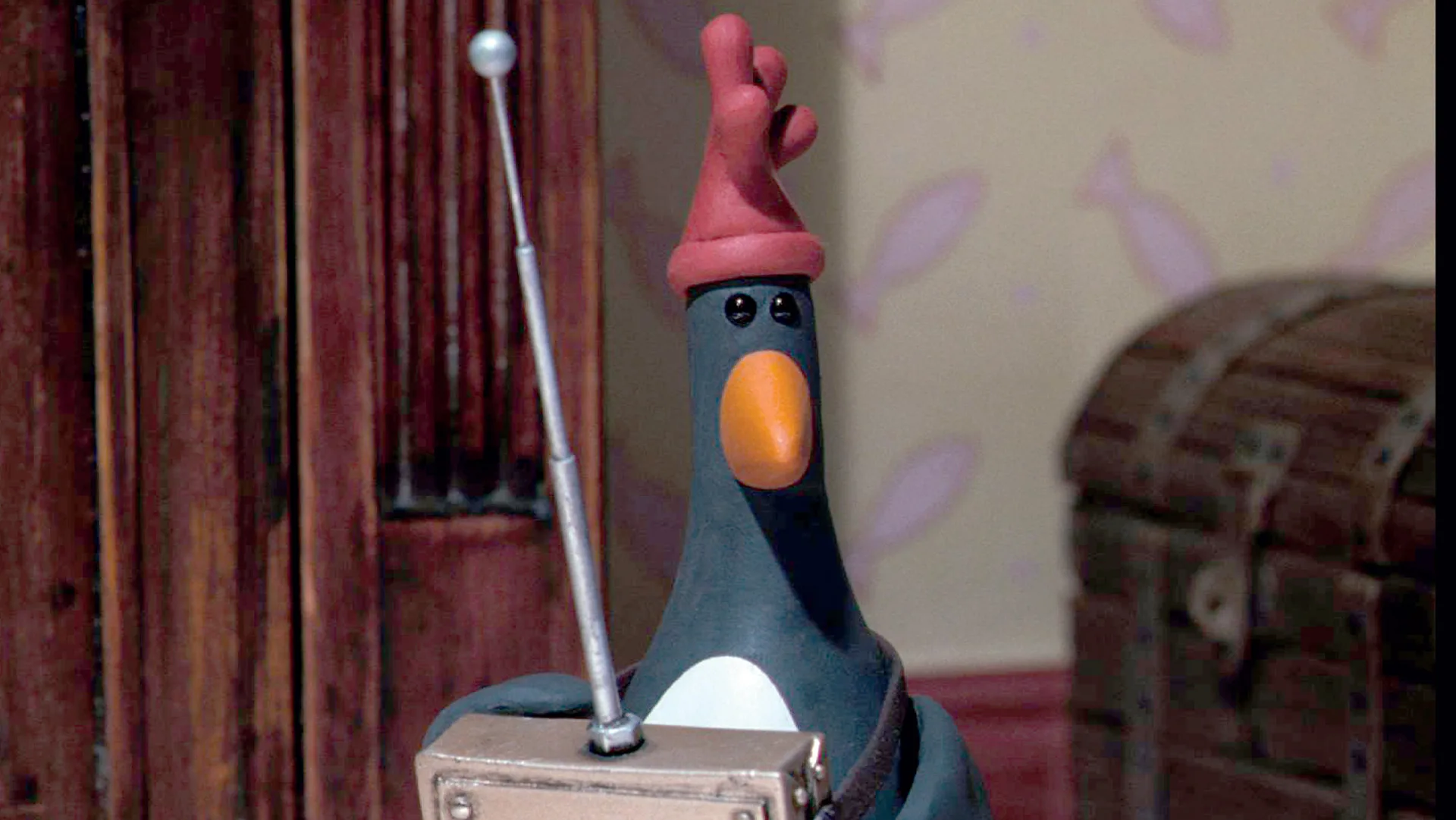 A still from Wallace and Gromit "The Wrong Trousers" showing the villain Feathers McGraw, a penguin with a rubber glove on his head made to look like a chicken, holding a remote control with an antennae in a room with a red curtain behind him and a wooden chest to his right.