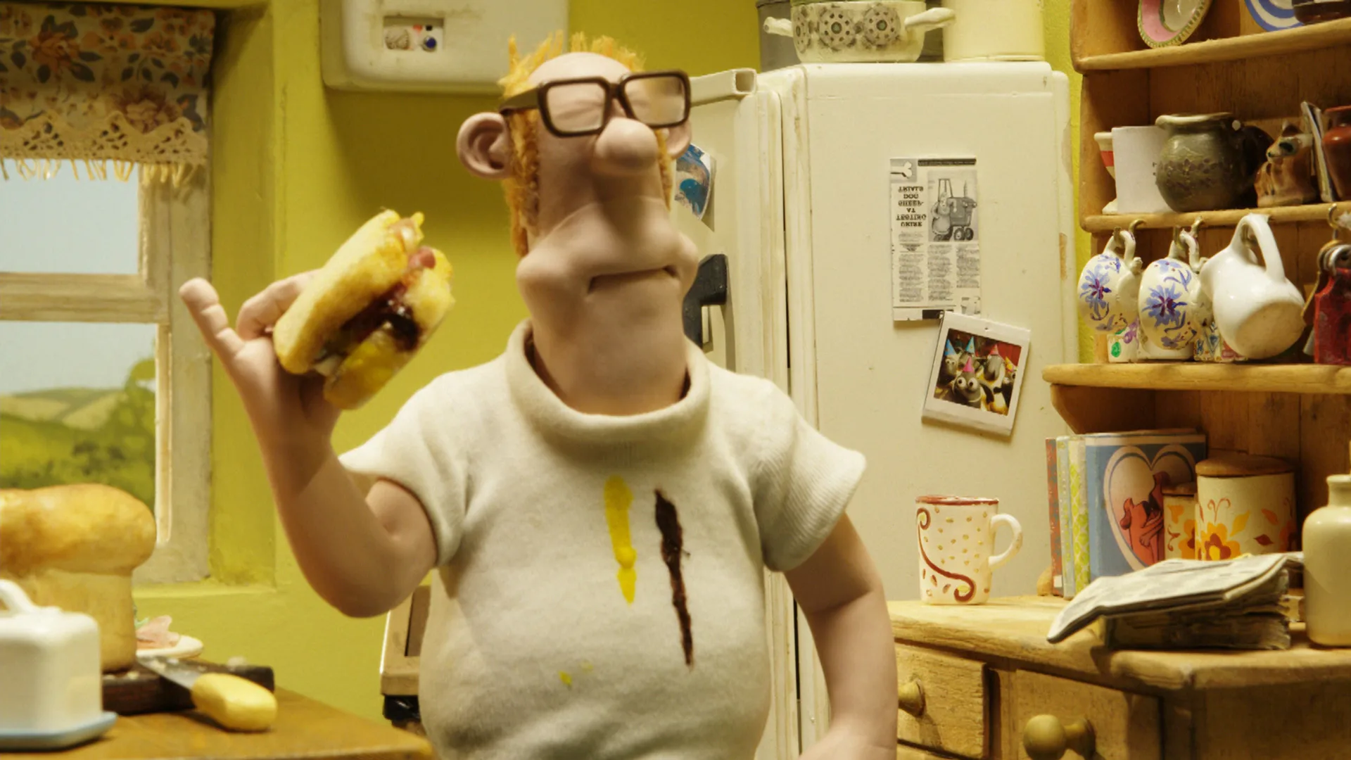 A still from Shaun the Sheep series showing The Farmer eating a sandwich in his kitchen wearing a white shirt covered in sauce stains from the sandwich.