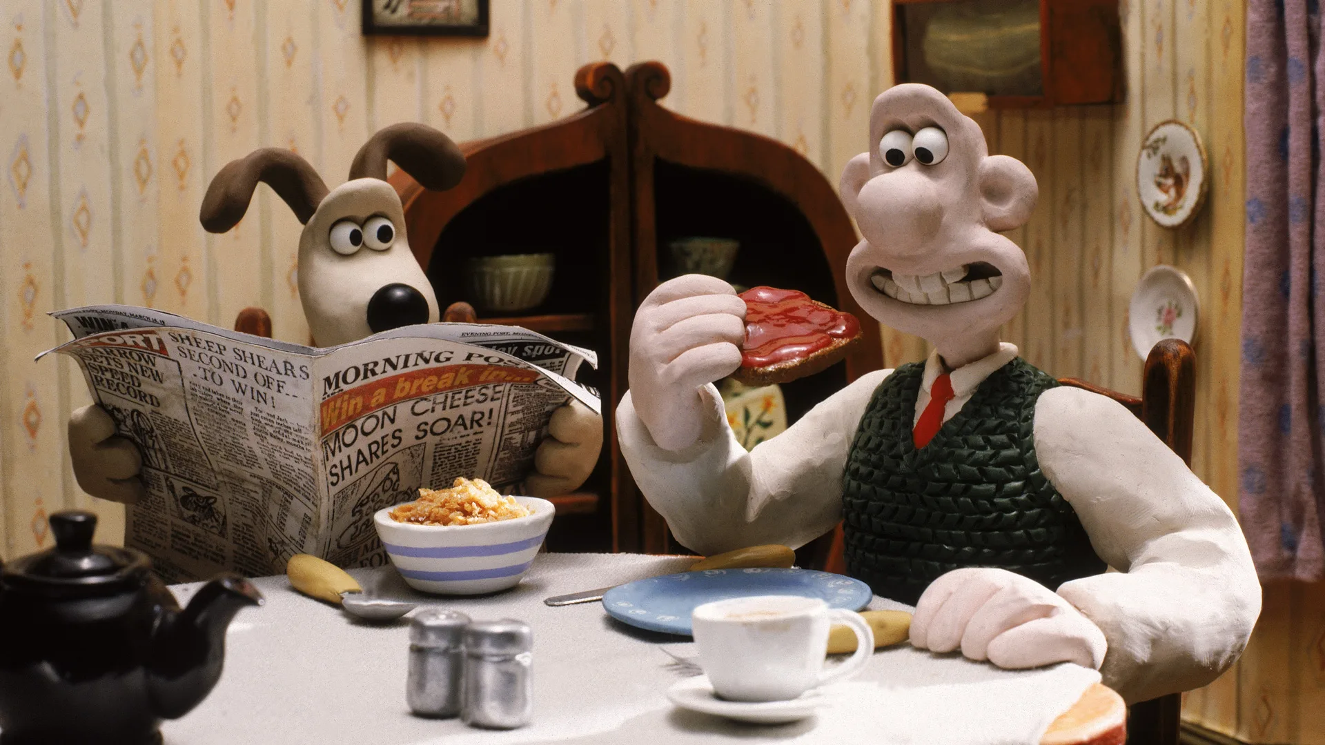 A still from Wallace and Gromit "The Wrong Trousers" showing the two sat at the breakfast table - Wallace is about to eat some jam on toast and Gromit is looking at him over his newspaper. There is a cup and bowl on the table and salt and pepper shakers. The background shows a wooden cabinet and yellow wallpaper.