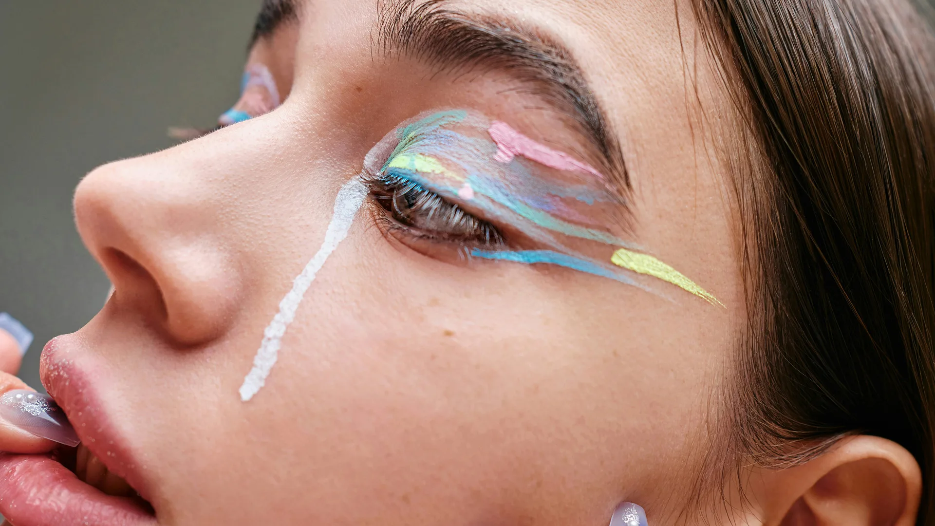 A close up photo of a girl with colourful eyeliner and white liner down her face like a tear. She is biting her colourful nails.