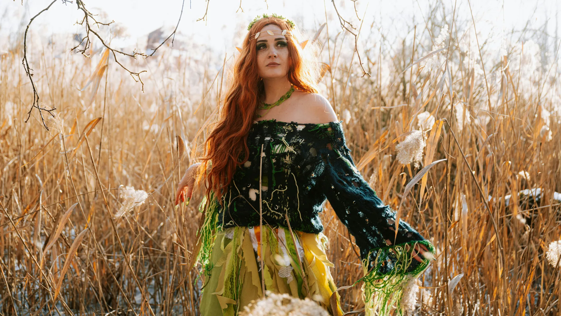A photo of a woman with red hair stood in a grassy field wearing a green ethereal outfit, arm stretched out with a flower crown on her head against a golden sunlit day.
