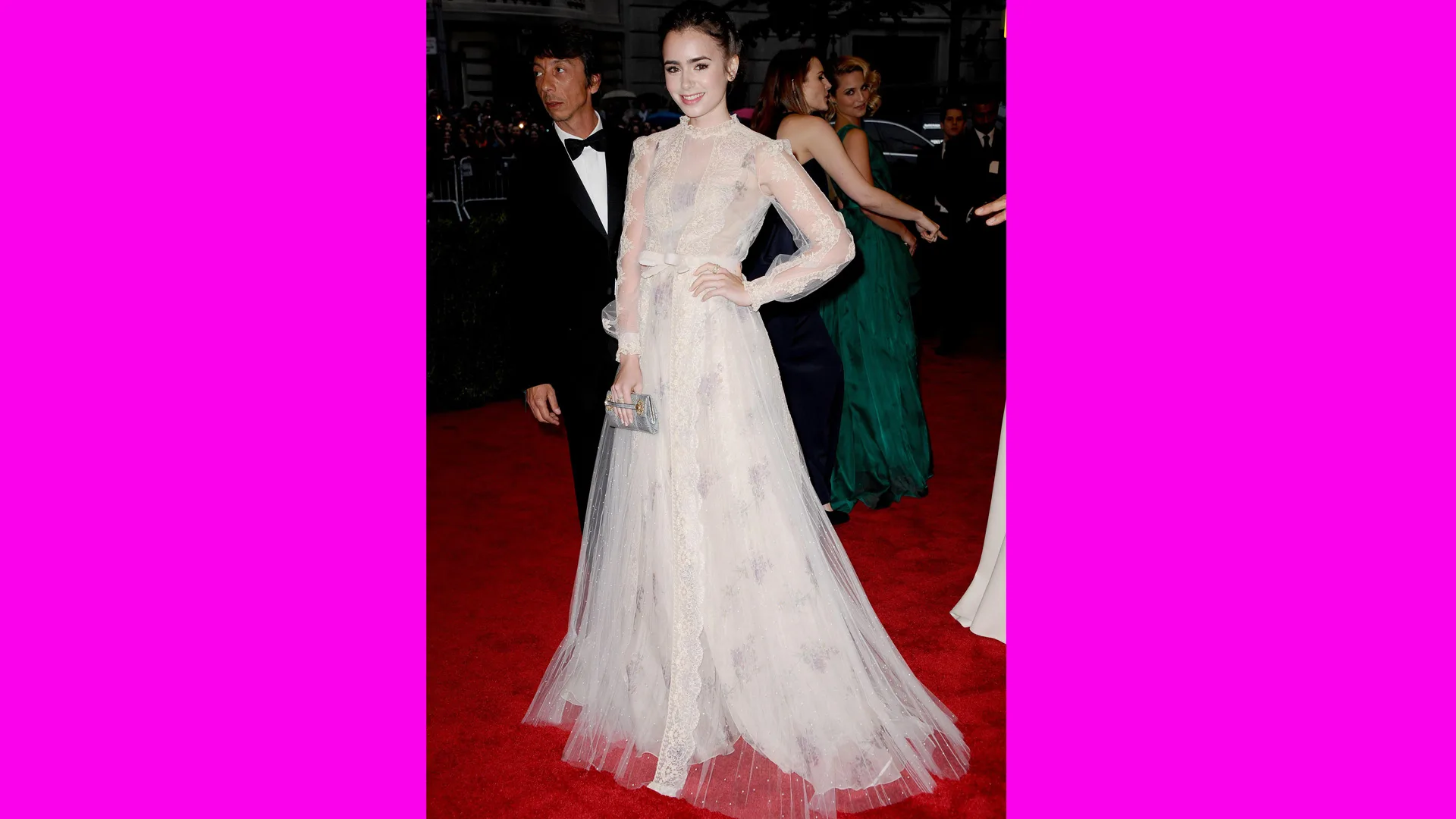 A photo of Lily Collins stood in a white lace Schiaparelli dress on the red carpet with people behind her.
