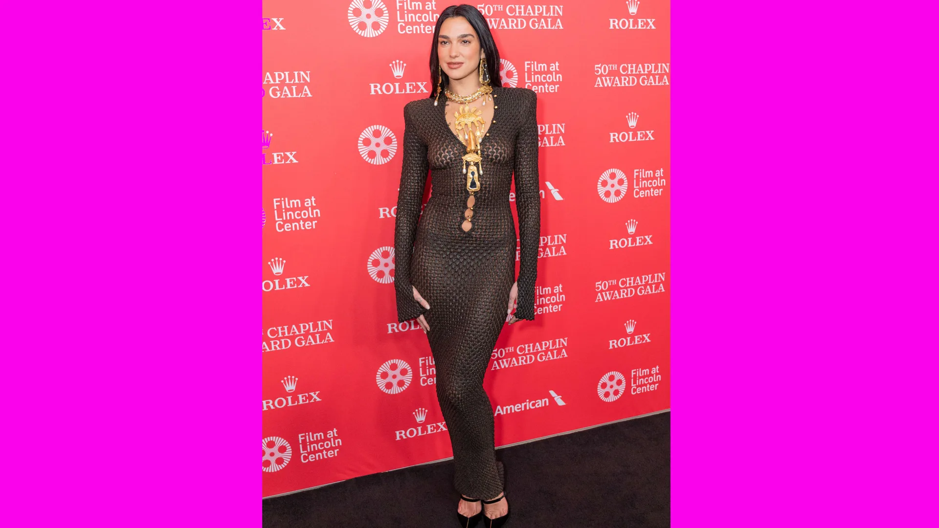 A photo of Dua Lipa stood on the red carpet wearing a black mesh bodysuit by Schiaparelli. She has a red background behind her with brand names.