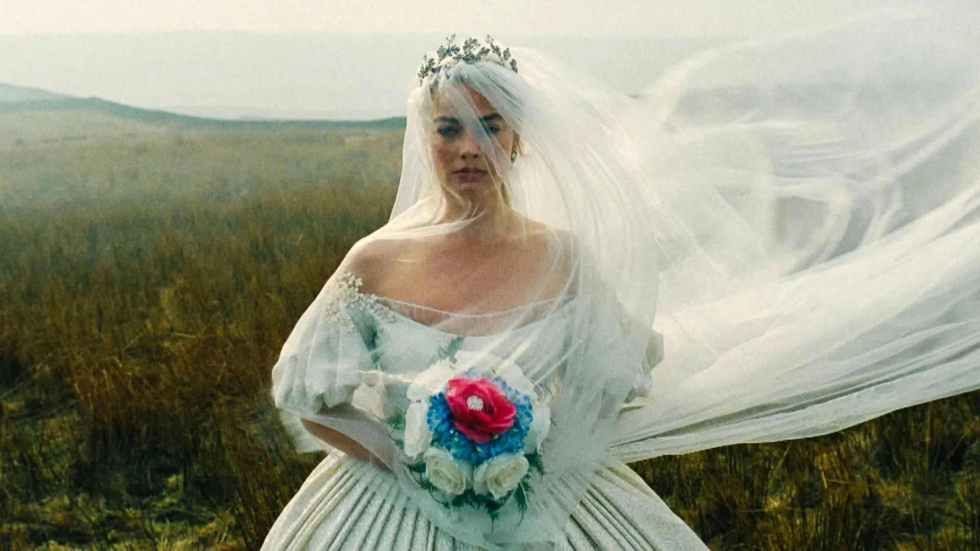 A still from Wuthering Heights (2026) showing Cathy in a white wedding gown holding a colourful bouquet - her veil is covering her face and blowing in the wind with the misty moors behind her.