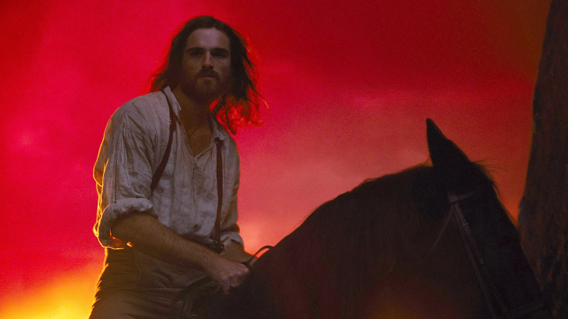 A still from Wuthering Heights (2026) showing Jacob Elordi as Heathcliff on a horseback with a red sunset behind him.