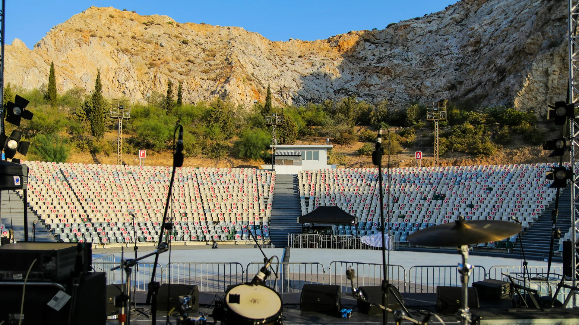 A photo of a stage with musical instruments including a drumkkit set up in an open air arena with seating and mountains and blue sky behind.