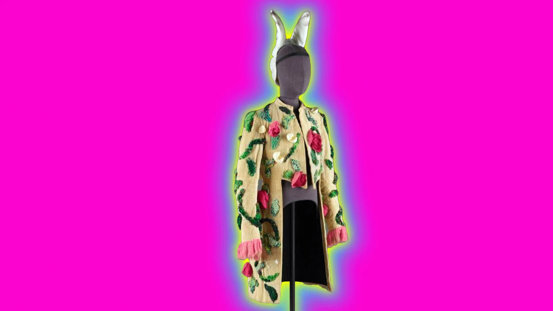A coat by Cecil Beaton in beige wool corduroy with applique flowers and stems climbing up the coat. It is hanged on a mannequin with rabbit ears on the head. The object is set against a neon pink background with a blue and yellow halo.