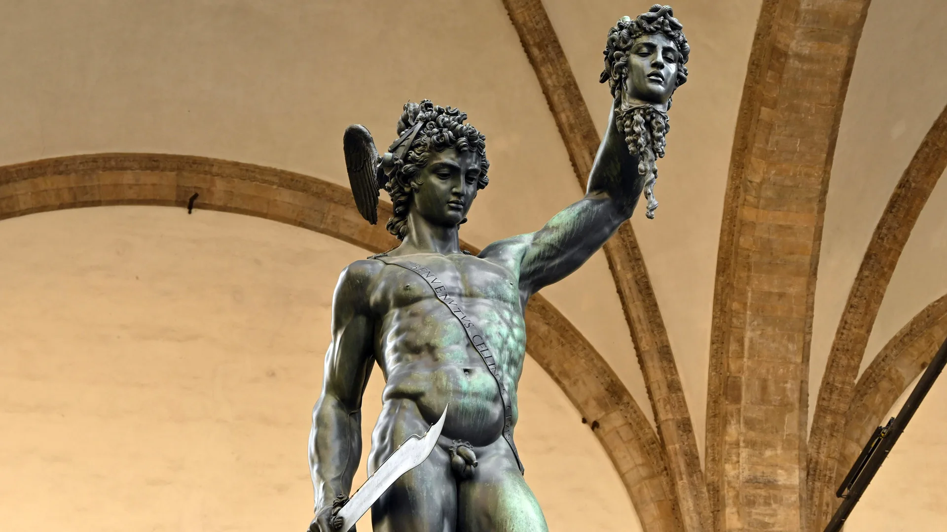 A photo of Perseus holding up the head of Medusa with a sword in his right hand. He is stood against an archway ceiling.