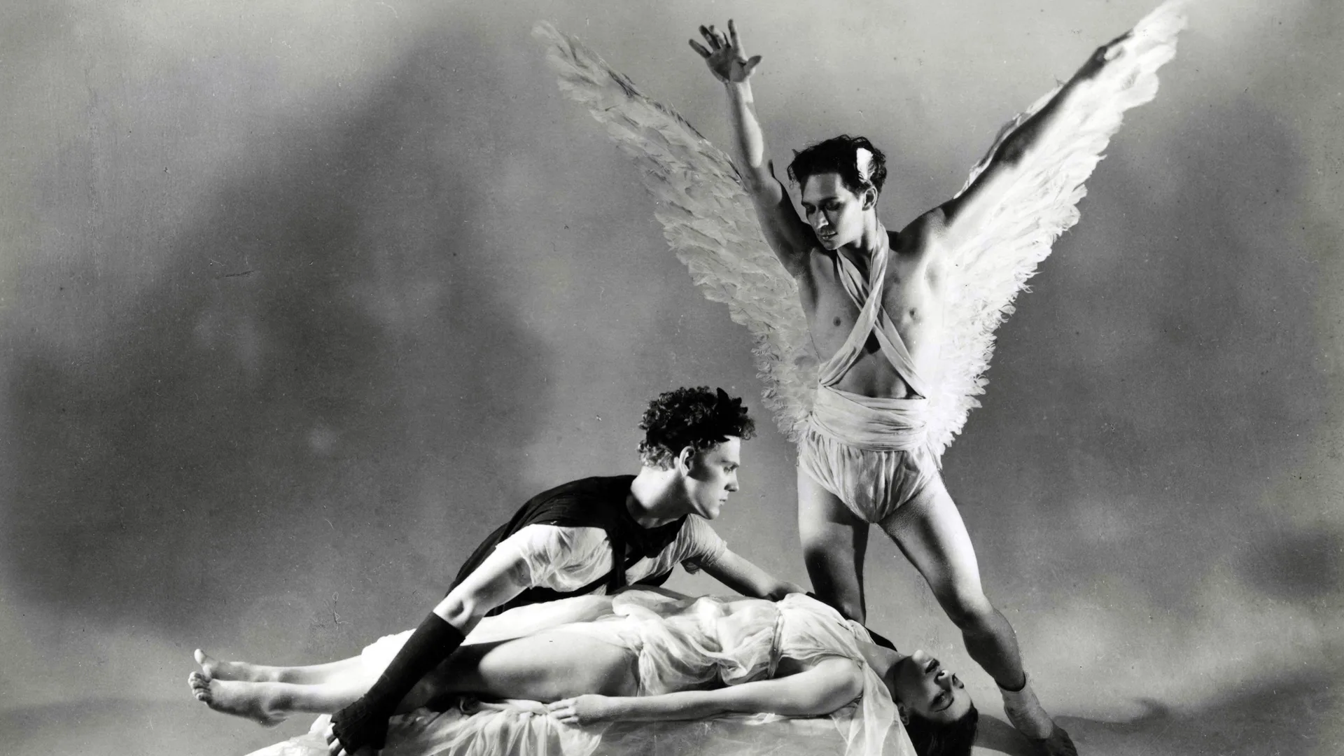A photo from Jerome Robbins Dance Division, The New York Public Library. It shows a group shot of the ballet Orpheus and Eurydice with Eurydice lying motionless on a plinth and Orpheus leans over her body and a man dressed as an angel arms outstretched above looks over them. There are clouds behind them.