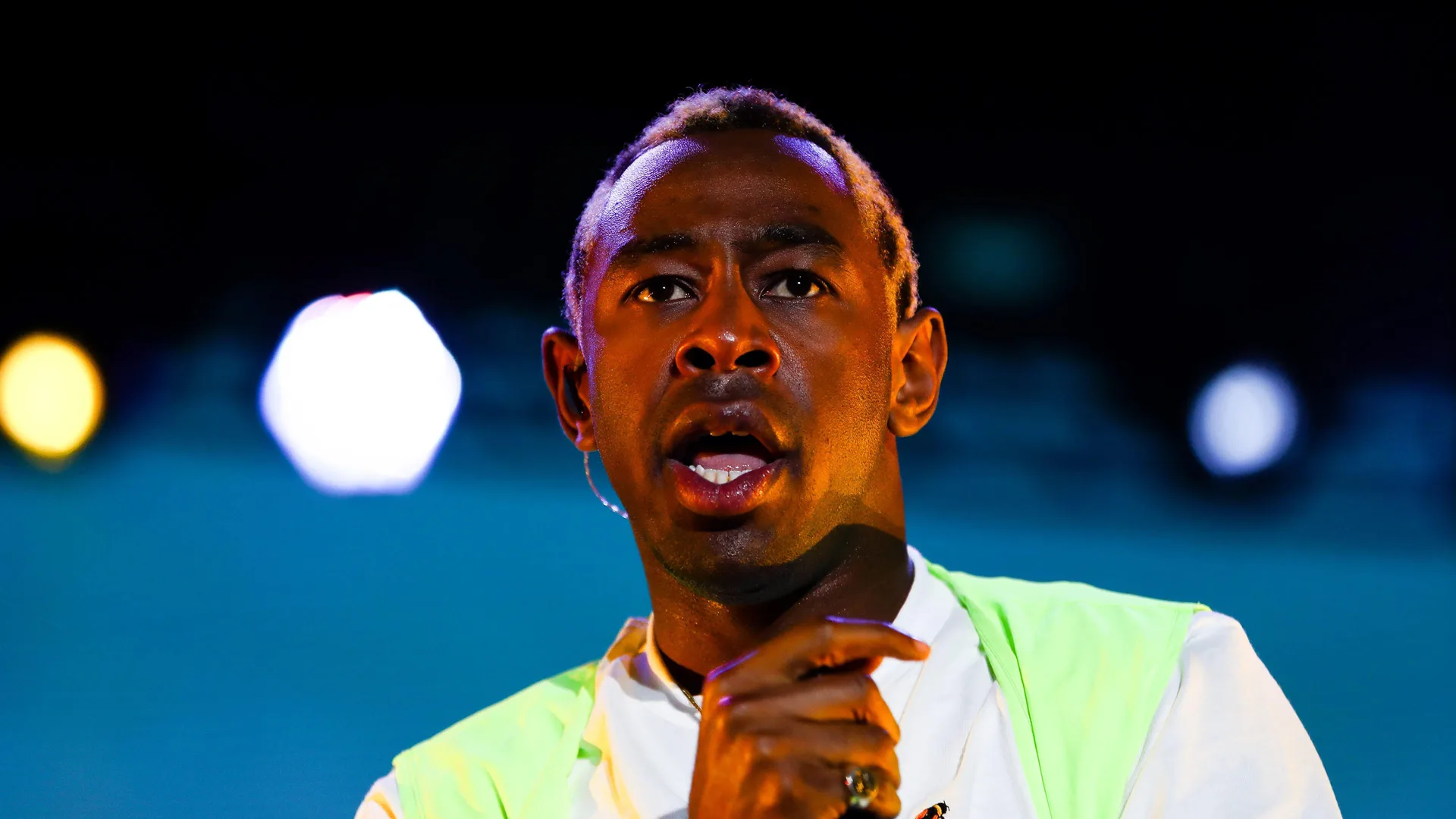 A photo of Tyler the Creator singing into a mic looking out to the distance against a blue light flared background.