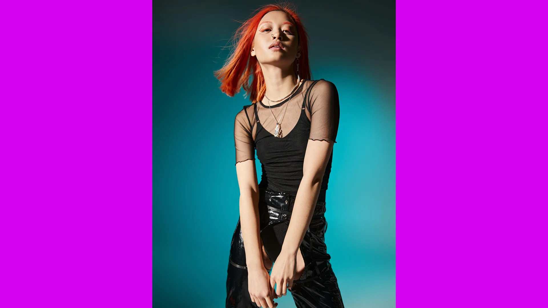 A photo of an asian model with red hair wearing a black sheer top with a black vest top over it and black jeans stood against a blue blackground with pink borders either side.