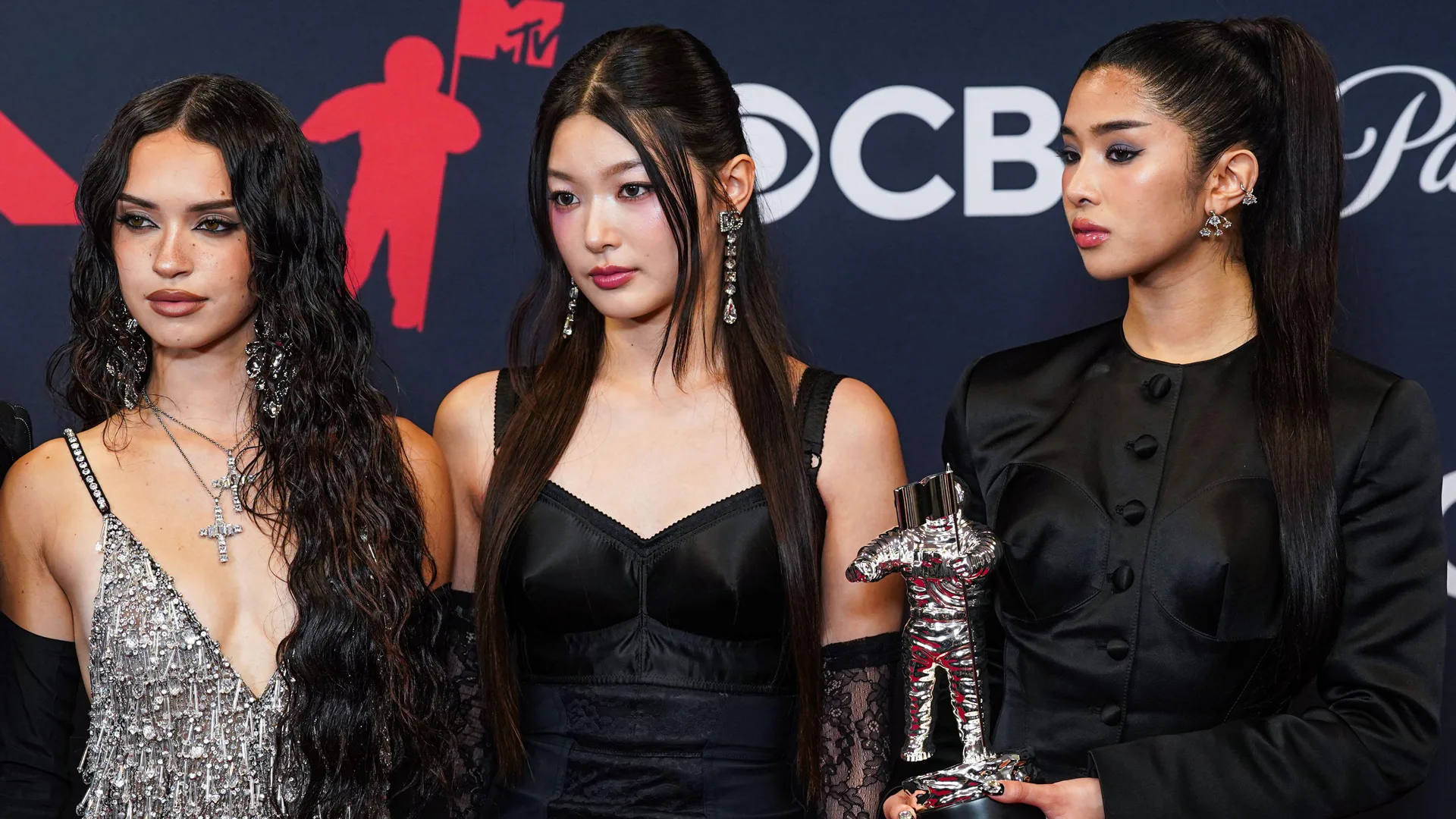 A photo of three members from the band KATSEYE stood at an awards show holding a silver award looking to the side to photographers.