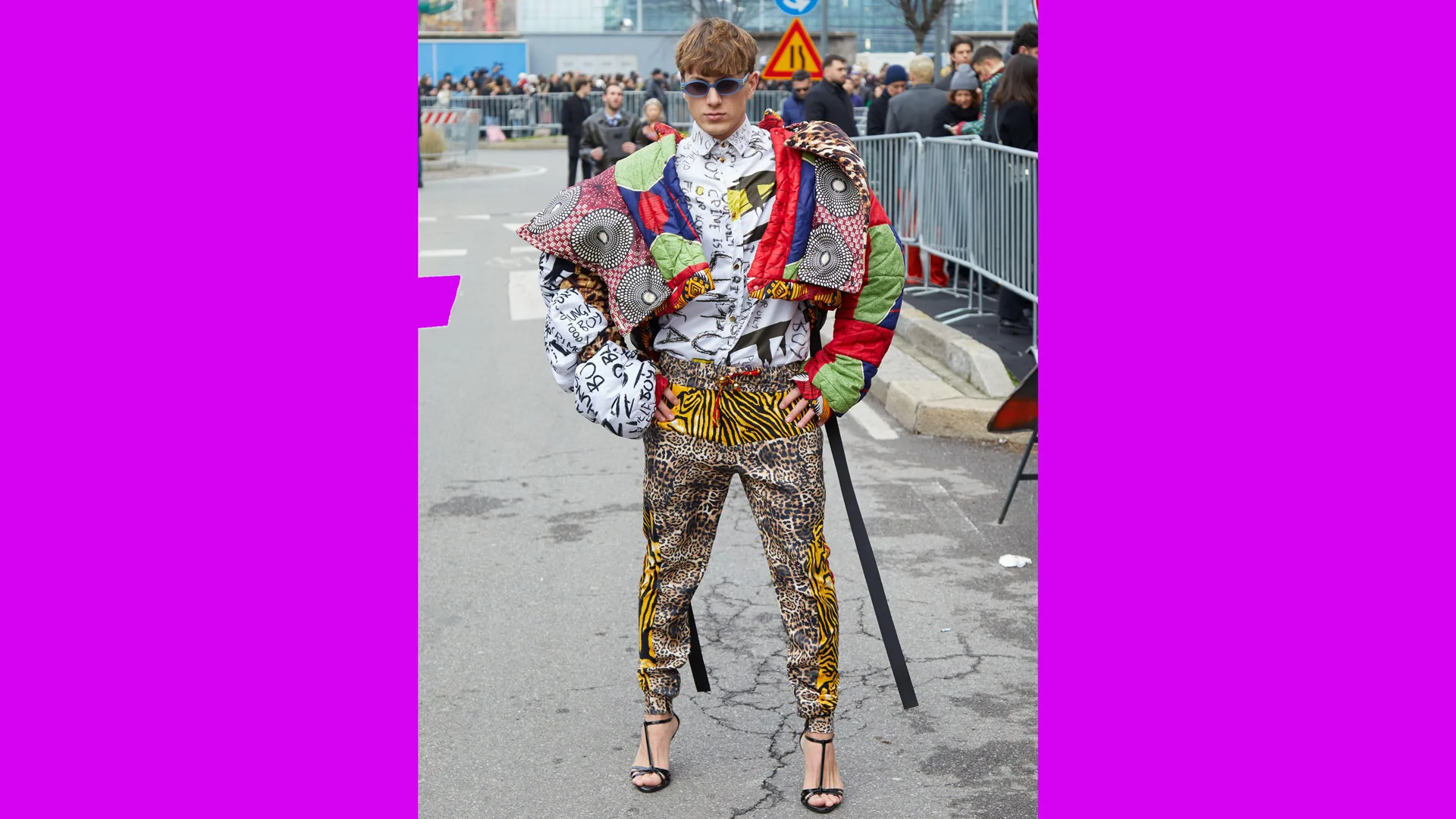 A photo of a man wearing an outfit of clashing colours and prints with heels stood at an event with people behind him.